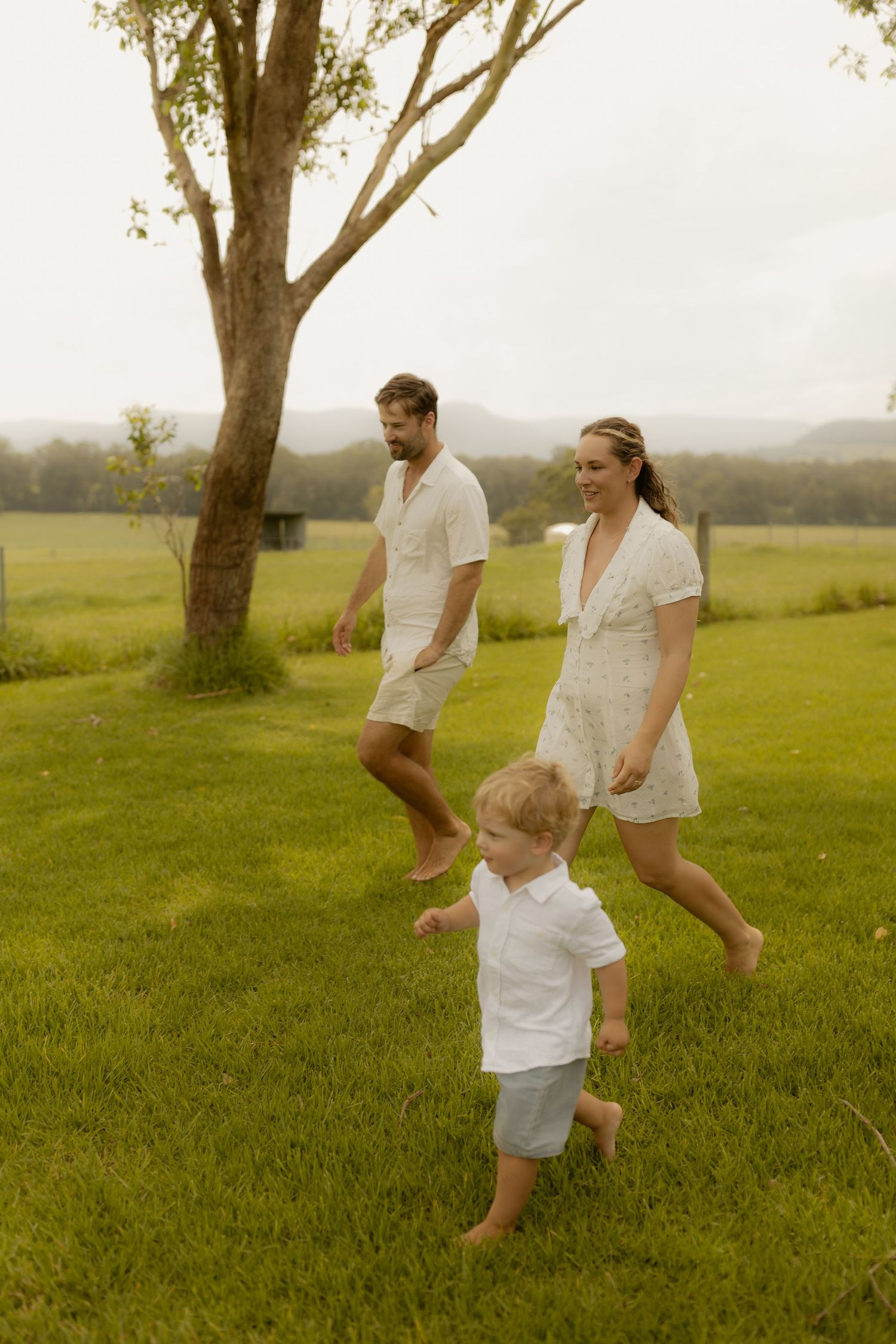 Family members in white clothing play and jump together on a lush green hillside.