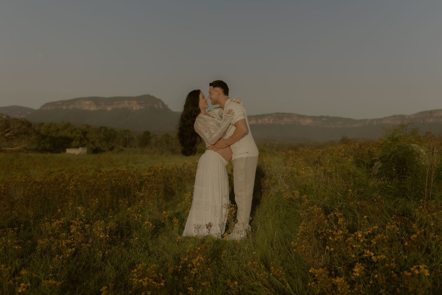 A couple shares an intimate moment in a field with mountains silhouetted against the dusky sky.