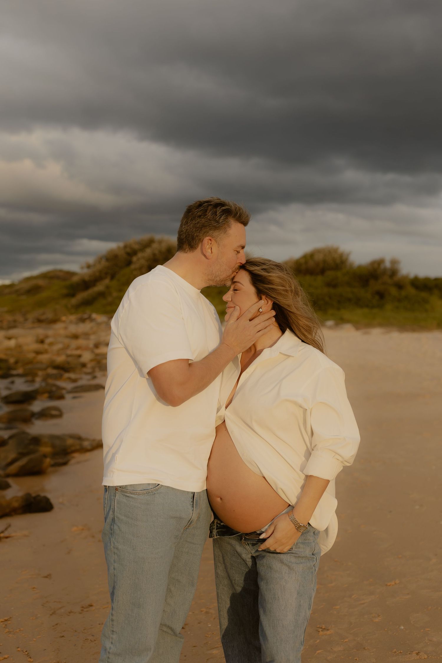 Couple sharing a tender moment on a beach, pregnant woman with exposed belly, man kissing her forehead under dramatic cloudy sky.