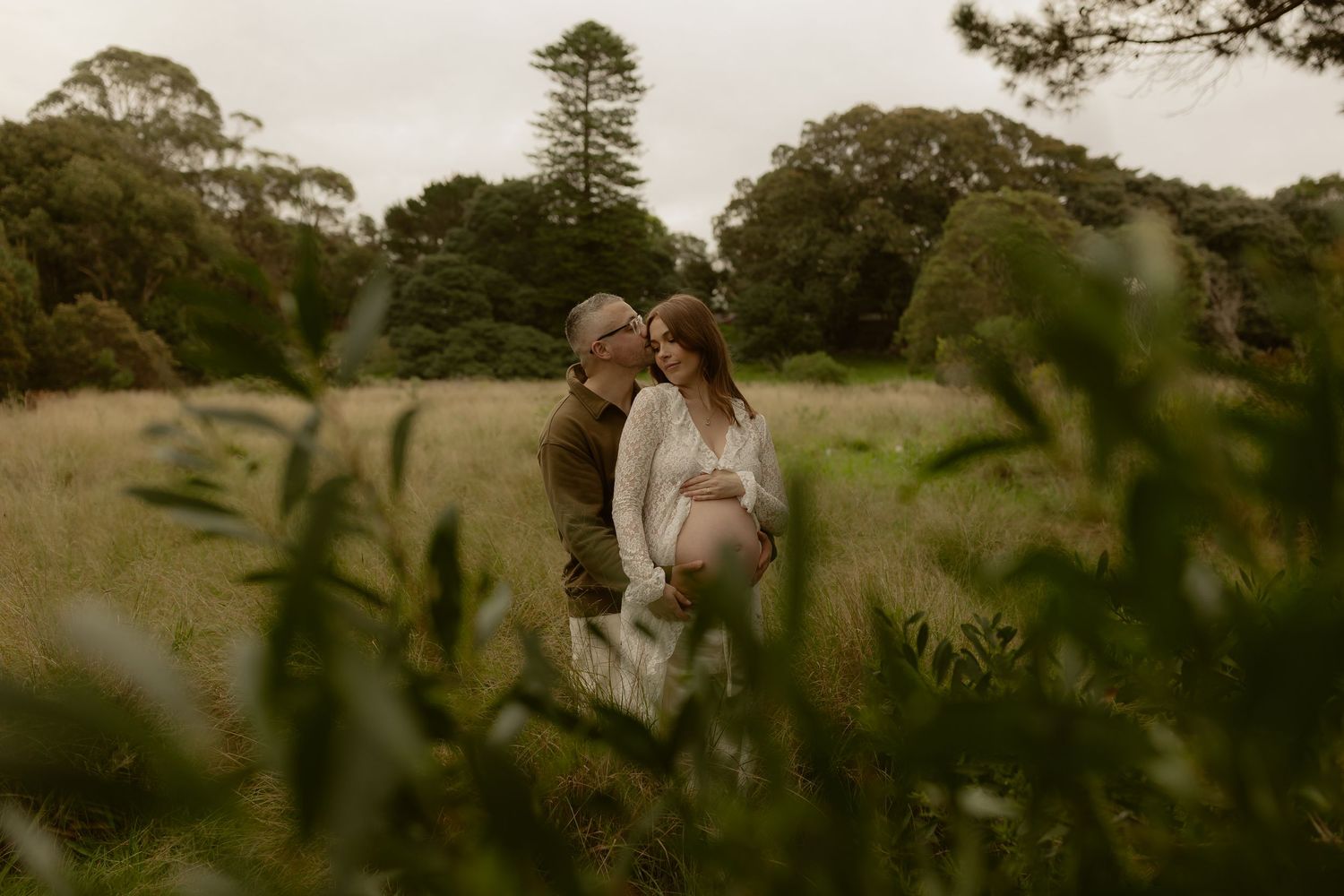 Couple embracing in lush green field, woman in white lace maternity dress, partner kissing her head.
