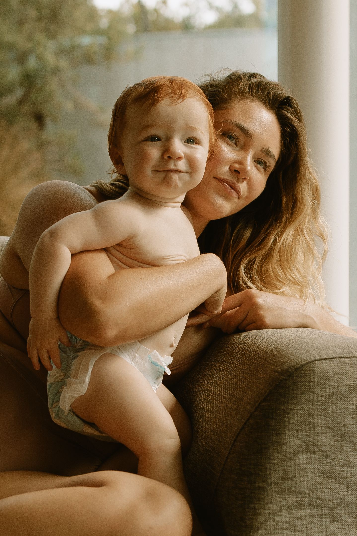A tender bonding moment between mother and baby sitting together on a gray couch in warm, natural lighting.