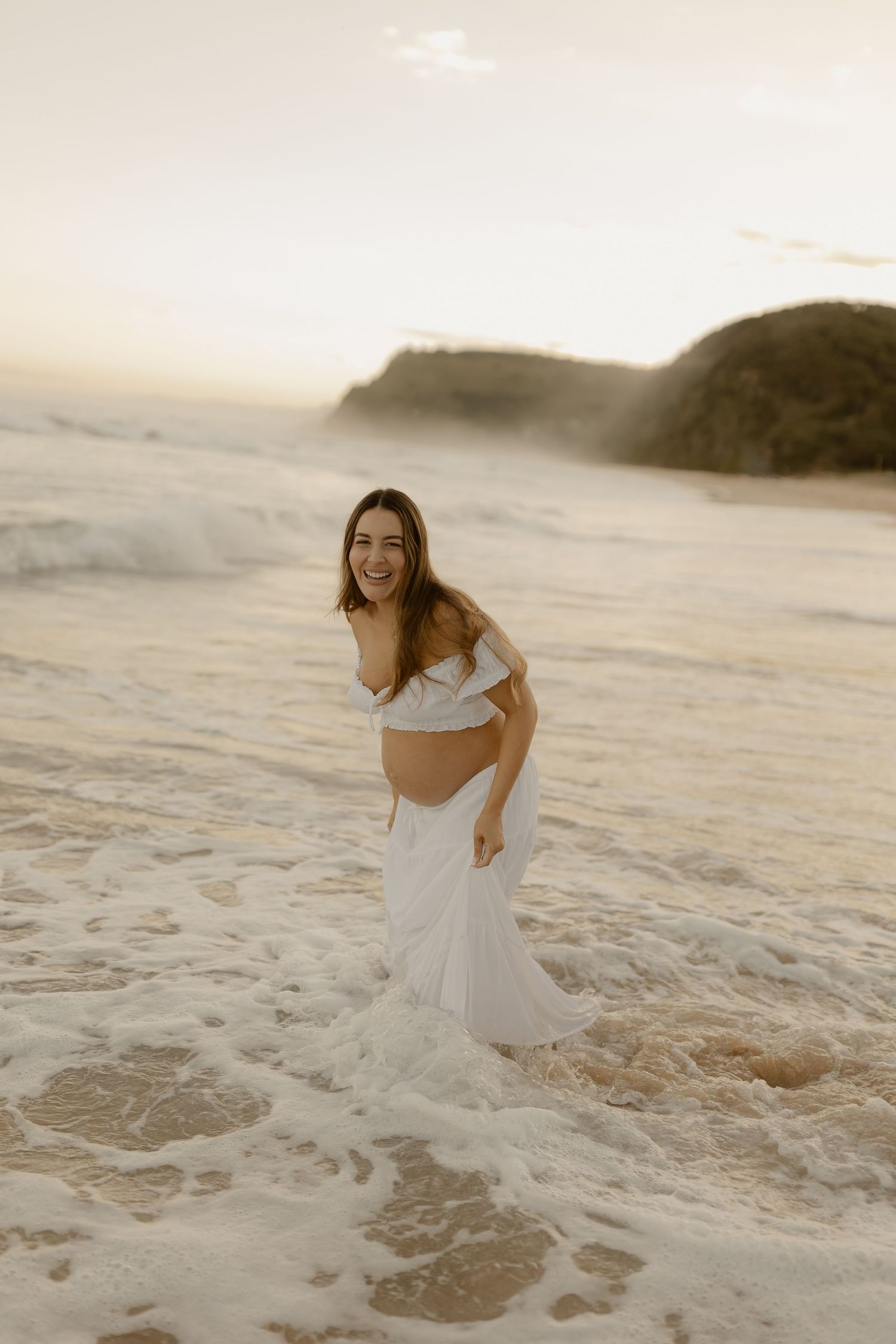 Woman in white dress playing joyfully in ocean waves at sunset on a sandy beach.