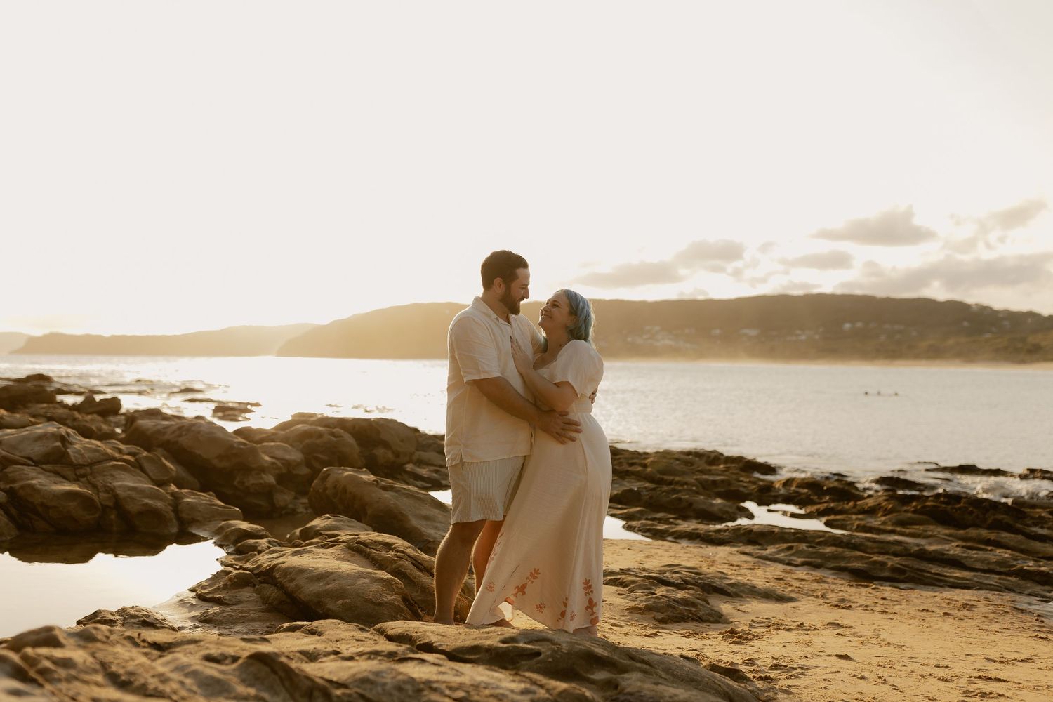 Couple embracing on rocky beach during sunset with ocean and mountains in background.