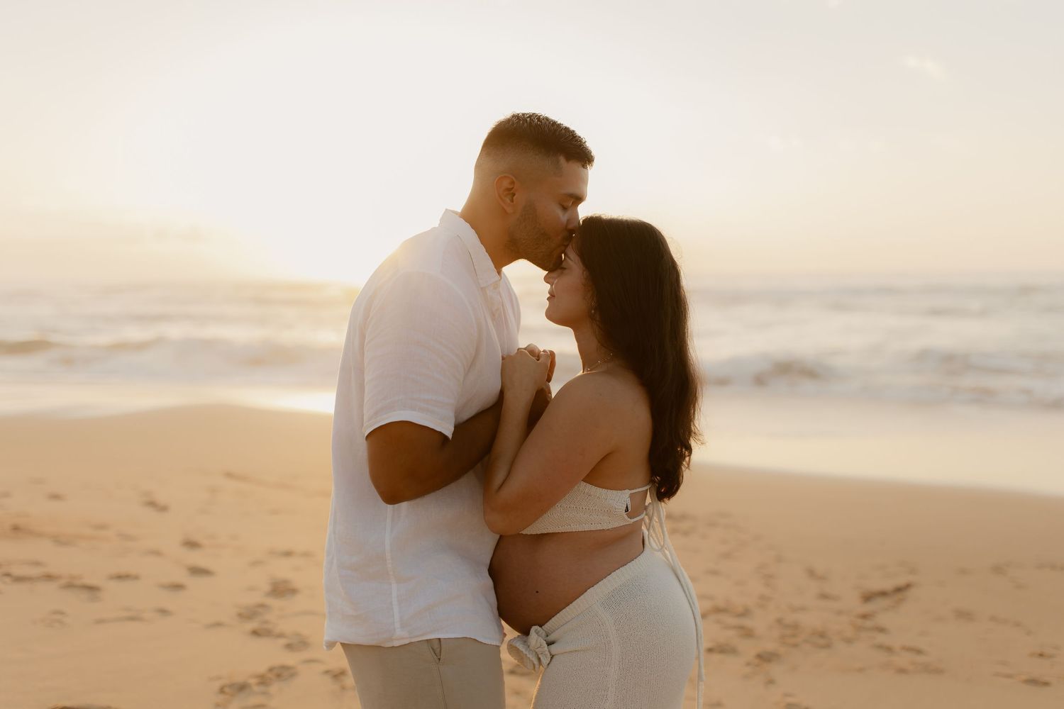 A couple shares an intimate moment at sunset on a sandy beach with ocean waves in the background.