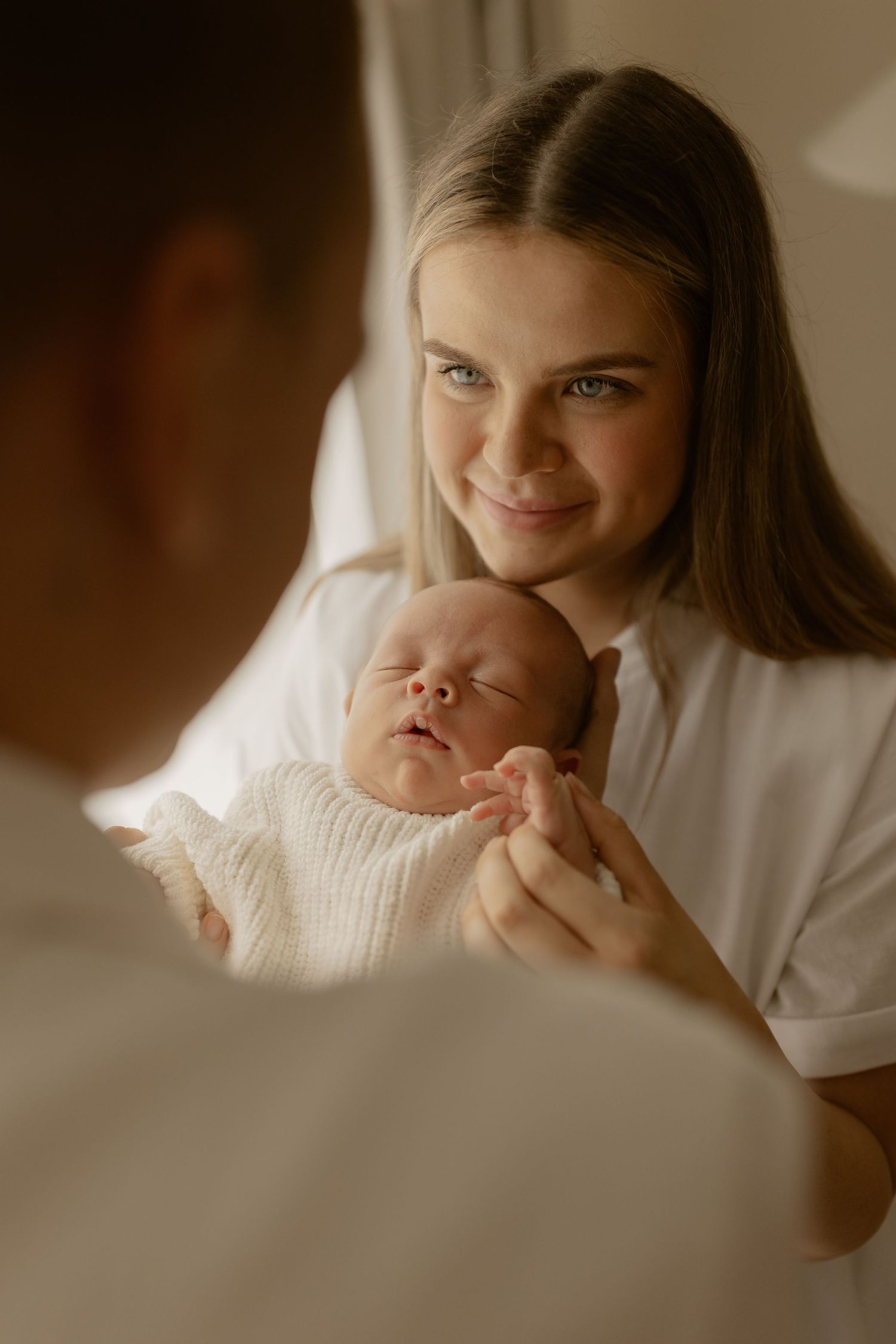 A sleeping newborn baby in a cream knit sweater being cradled while bathed in warm natural light.