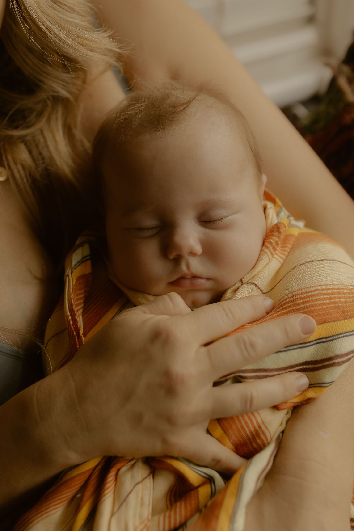A sleeping newborn baby wrapped in a striped orange blanket, cradled in a mother's arms.
