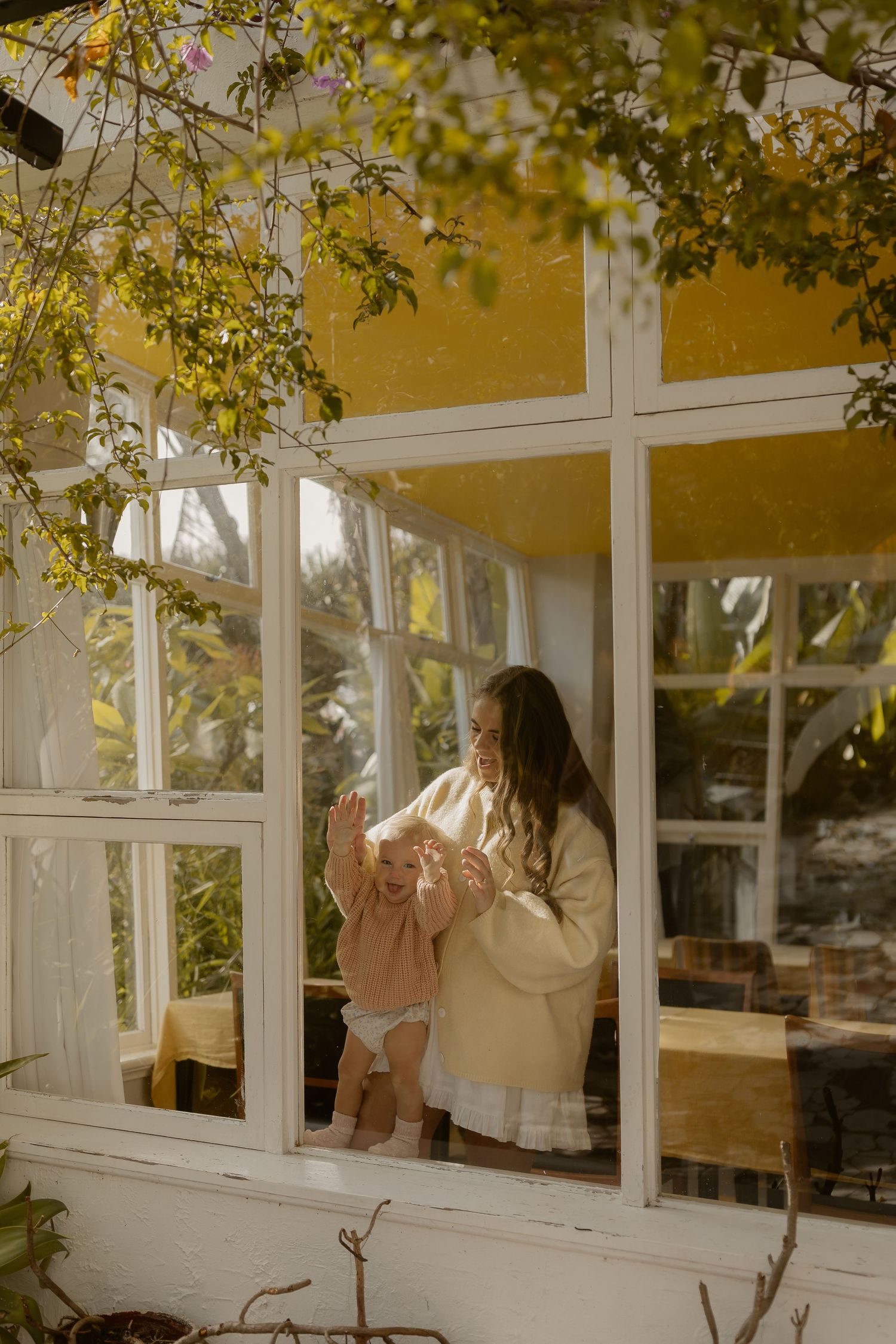 Mother and baby playing together by a sunlit greenhouse window surrounded by lush green foliage.