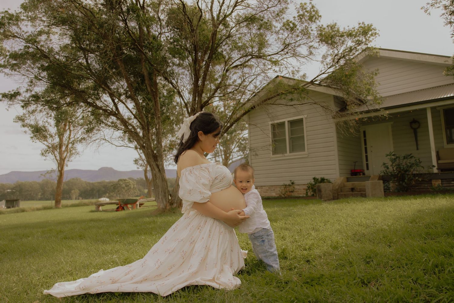 Pregnant woman in white dress plays with toddler son on green lawn outside a country home.