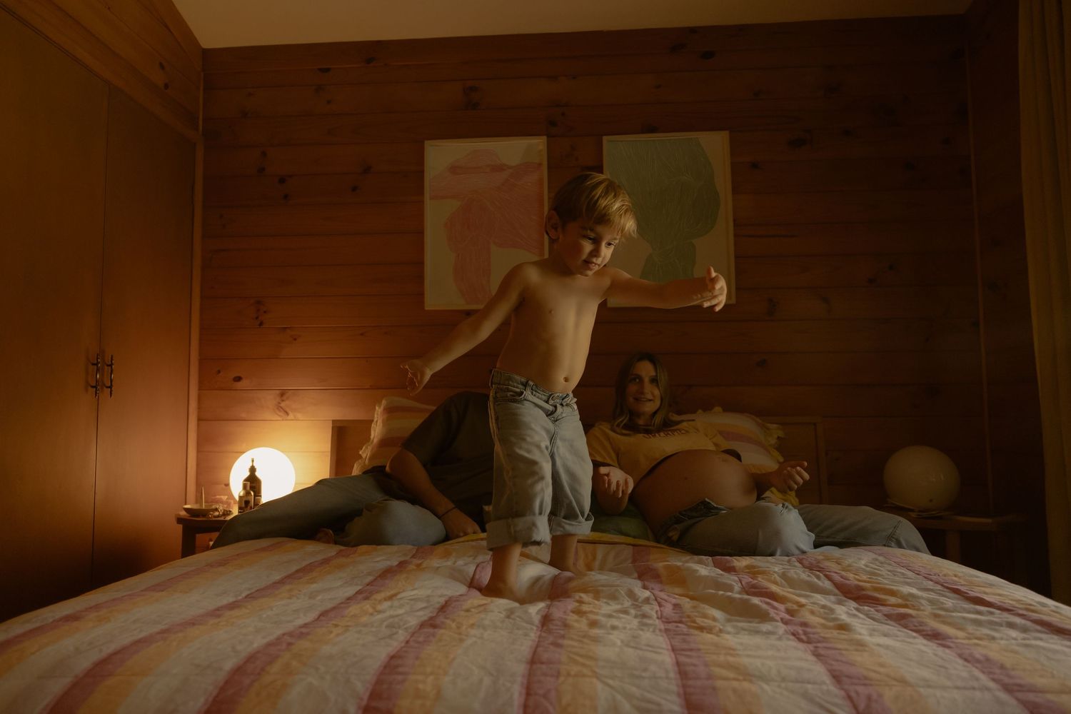 Child jumping on a bed in a cozy wooden cabin bedroom with warm lighting and an adult resting nearby.