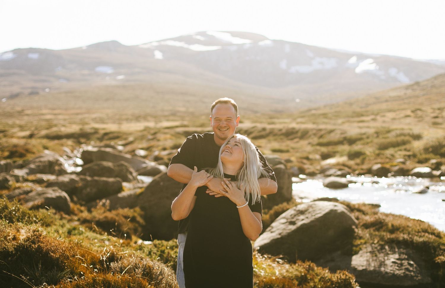 A hiker embraces companion among golden rocks in mountainous terrain during sunset with snow-capped peaks in background.