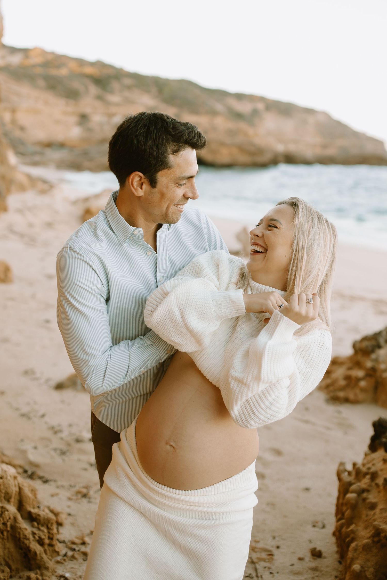 Expecting couple shares a joyful moment together on a beach during sunset with rocky coastline in background.