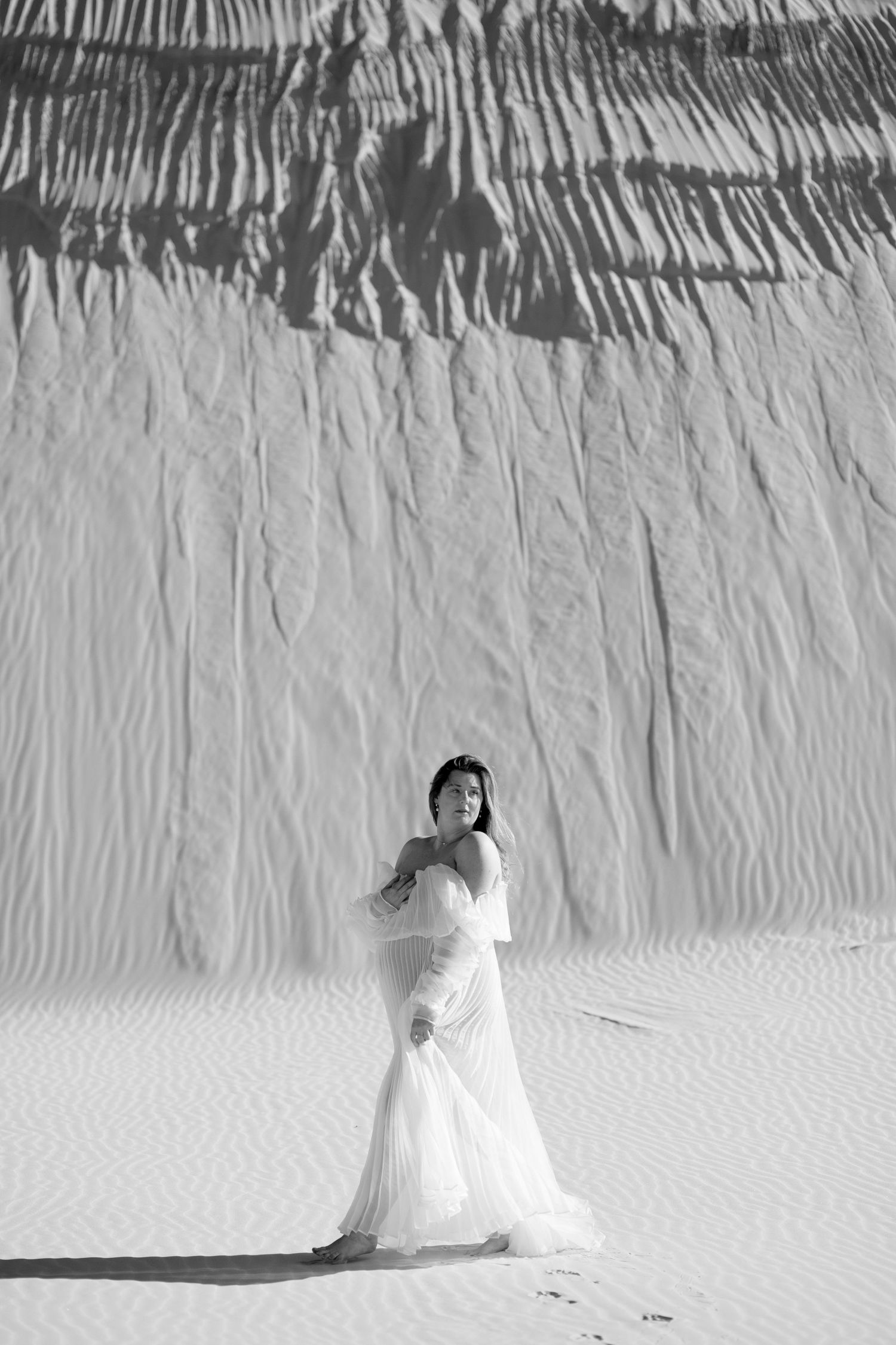Black and white photo of person in flowing white dress posing in front of textured backdrop creating dramatic contrast.