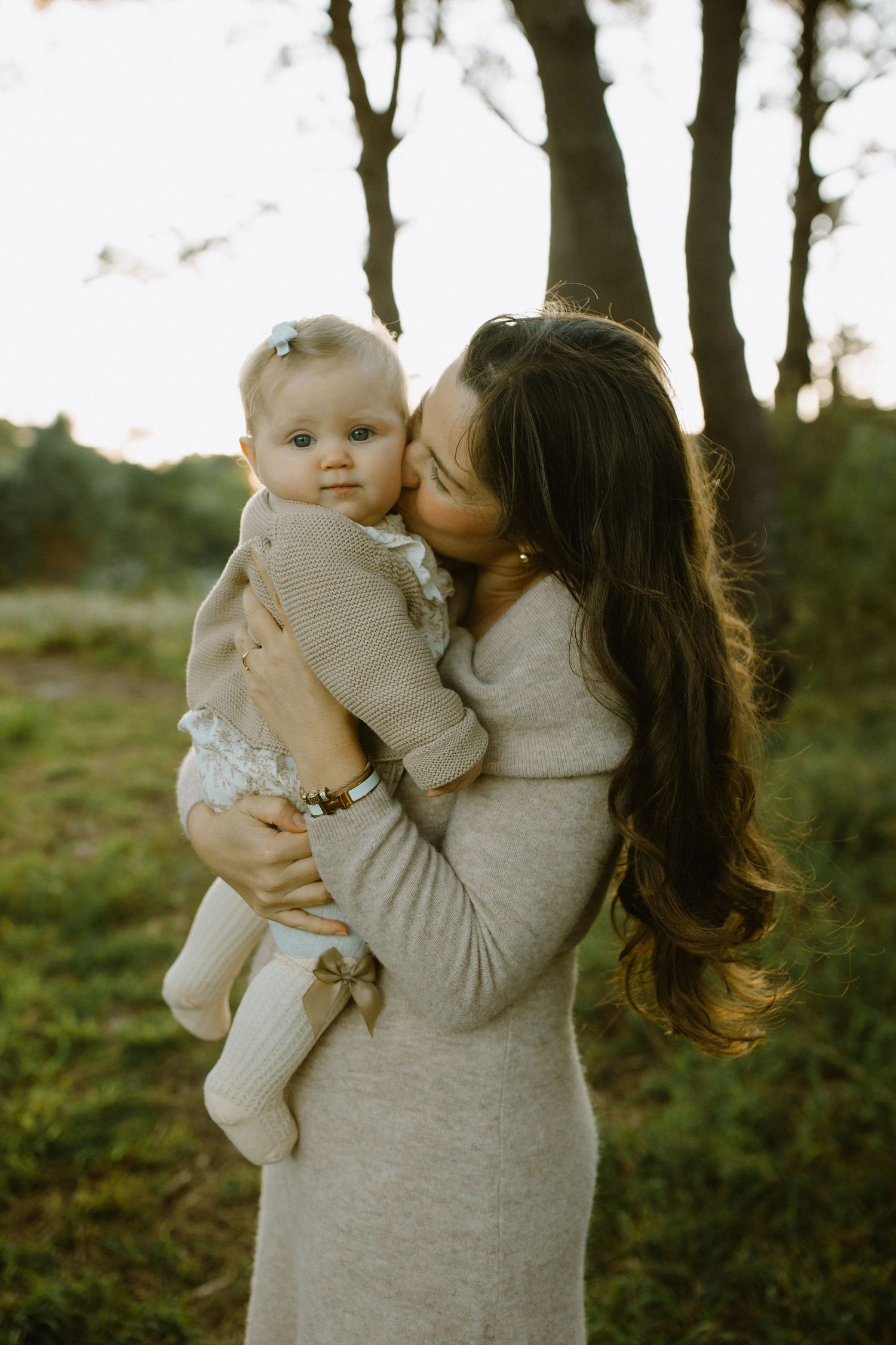 Mother in grey dress holds and kisses baby in light colored outfit against forest background.