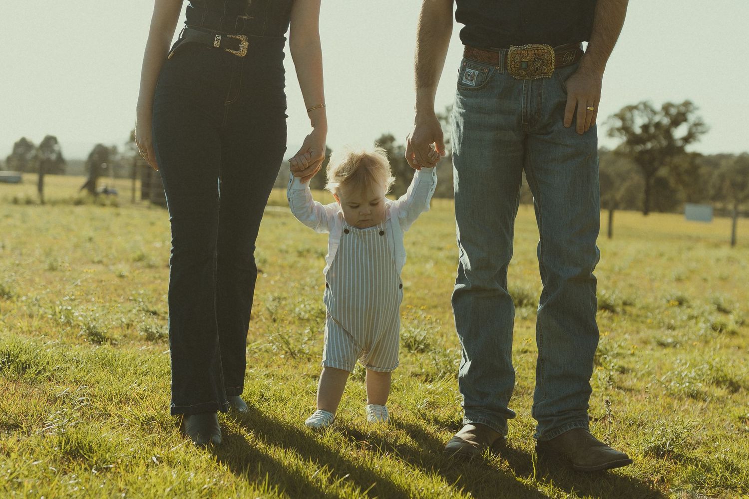 People's legs and feet shown standing together in a grassy field at sunset.