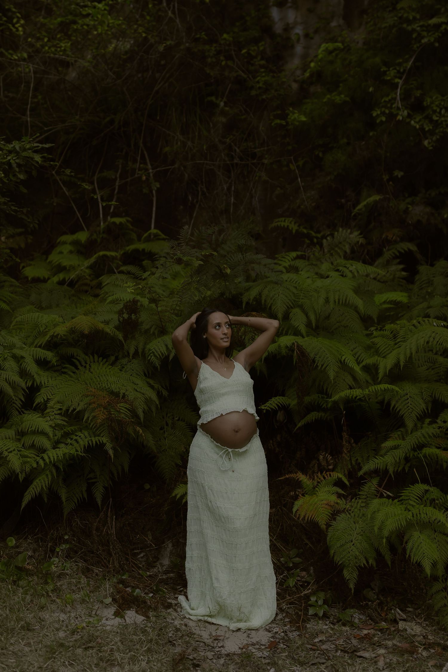 Pregnant woman in white outfit stands among ferns in dark forest, hands raised to hair.