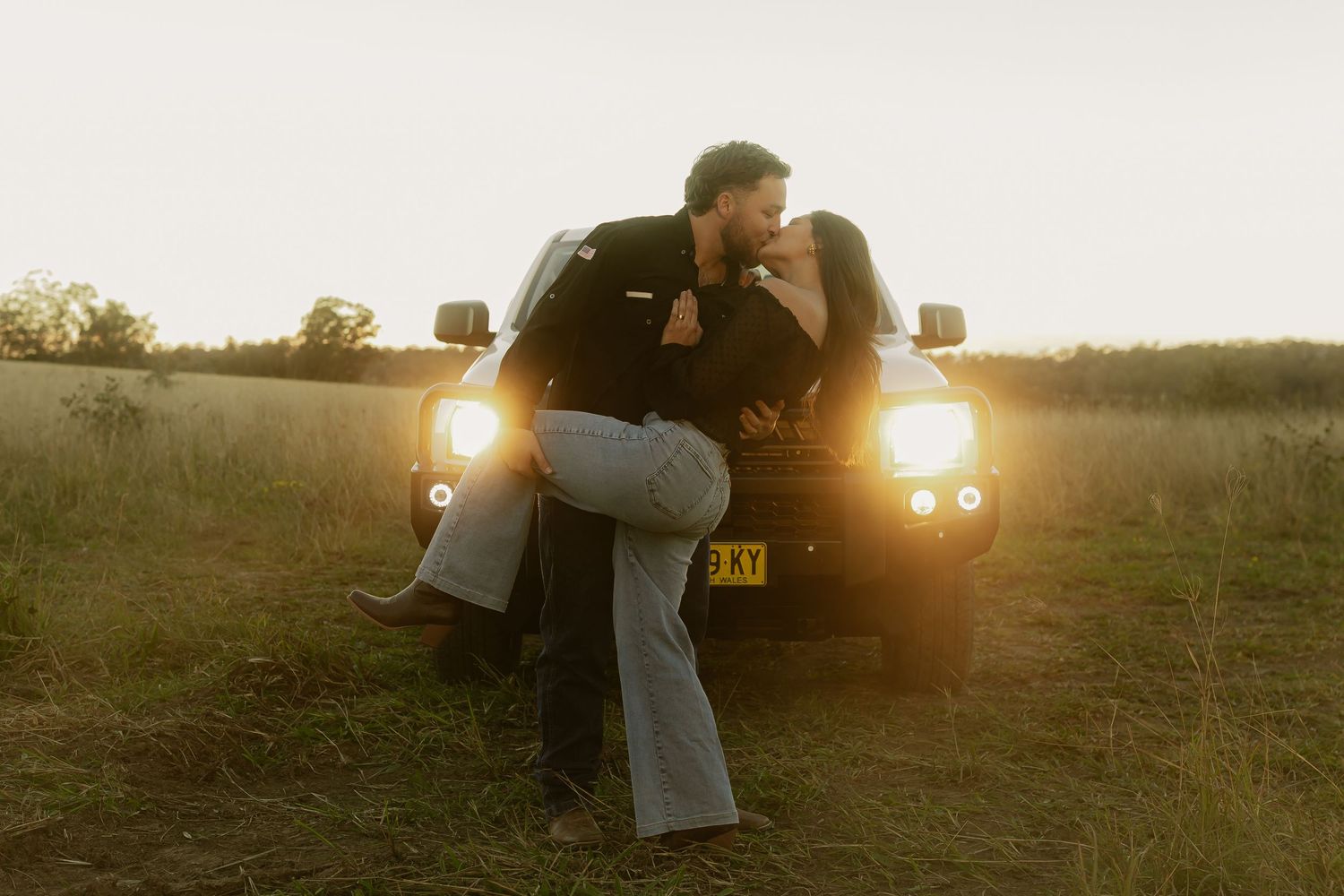 Couple sharing a romantic kiss leaning against a truck in a golden field at sunset.