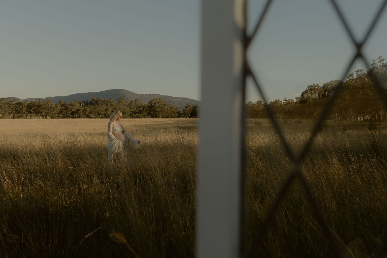 Person in white dress standing in golden field with mountain backdrop, viewed through fence at dusk.