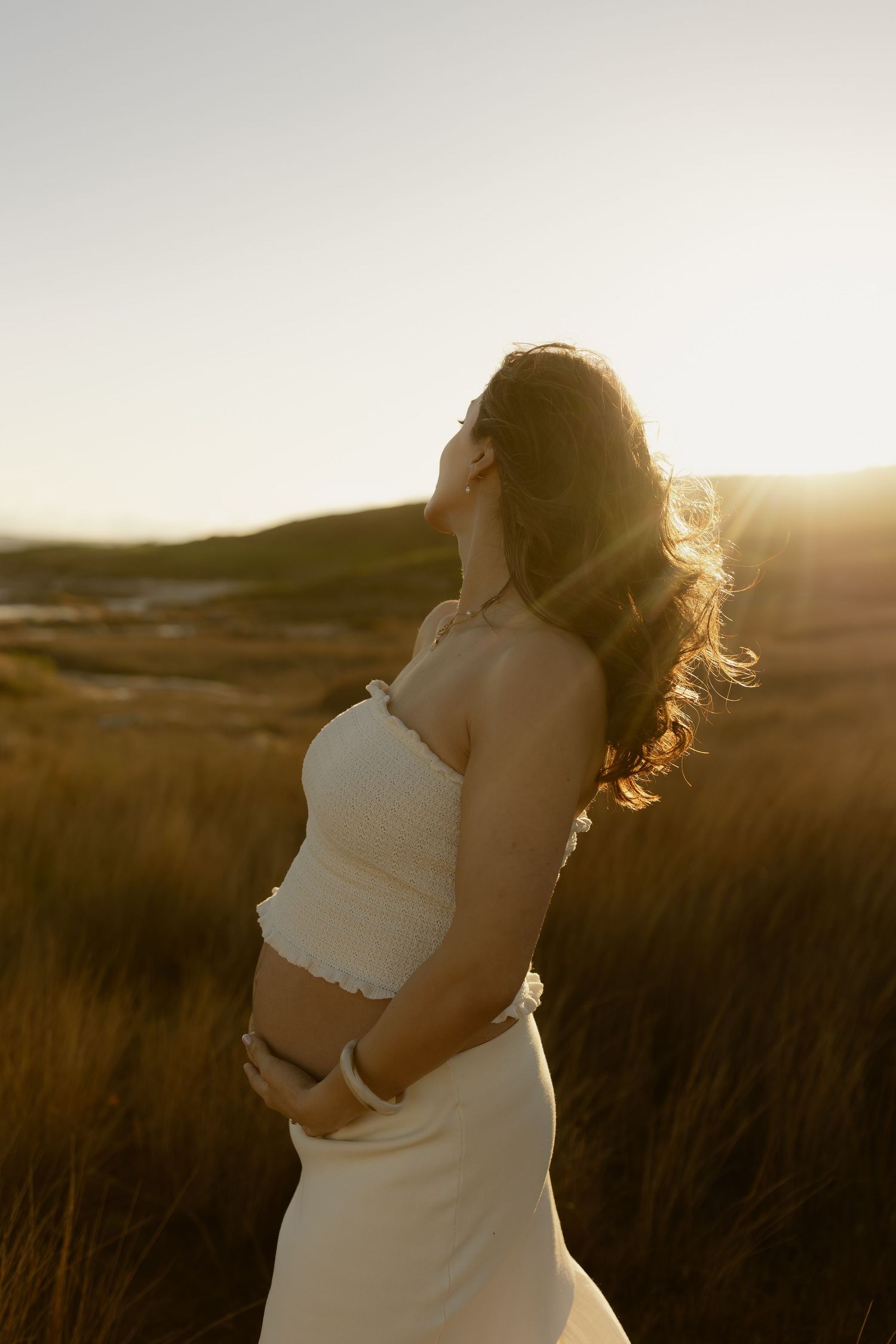 Pregnant woman in white dress holding baby bump at golden sunset in grassy field.