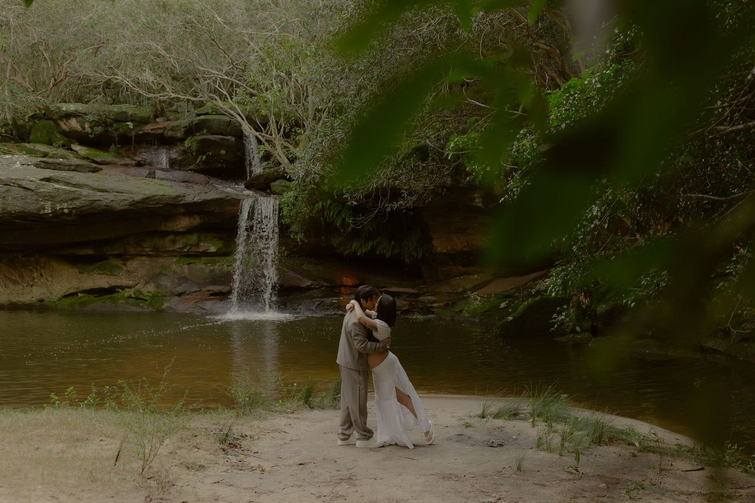 Couple embracing near a waterfall in a lush forest setting, bride in white gown, ideal for wedding photography inspiration.