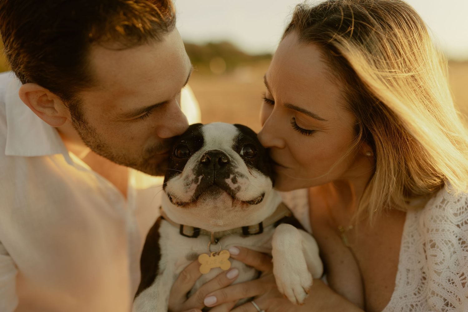 Couple kissing a black and white French Bulldog outdoors in golden hour light.