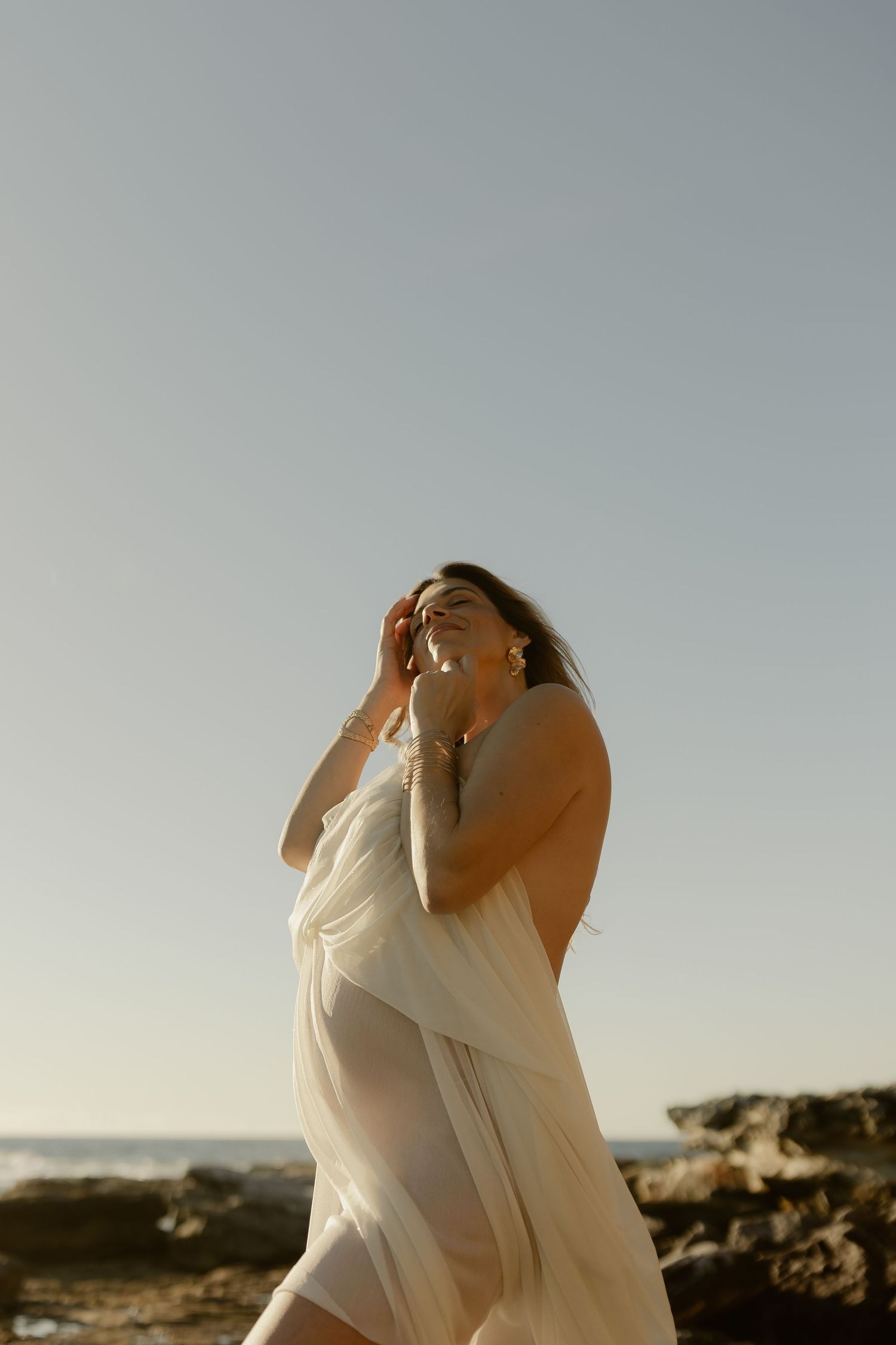 Woman in sheer white dress poses on rocky beach at golden hour, hair blowing in the breeze.