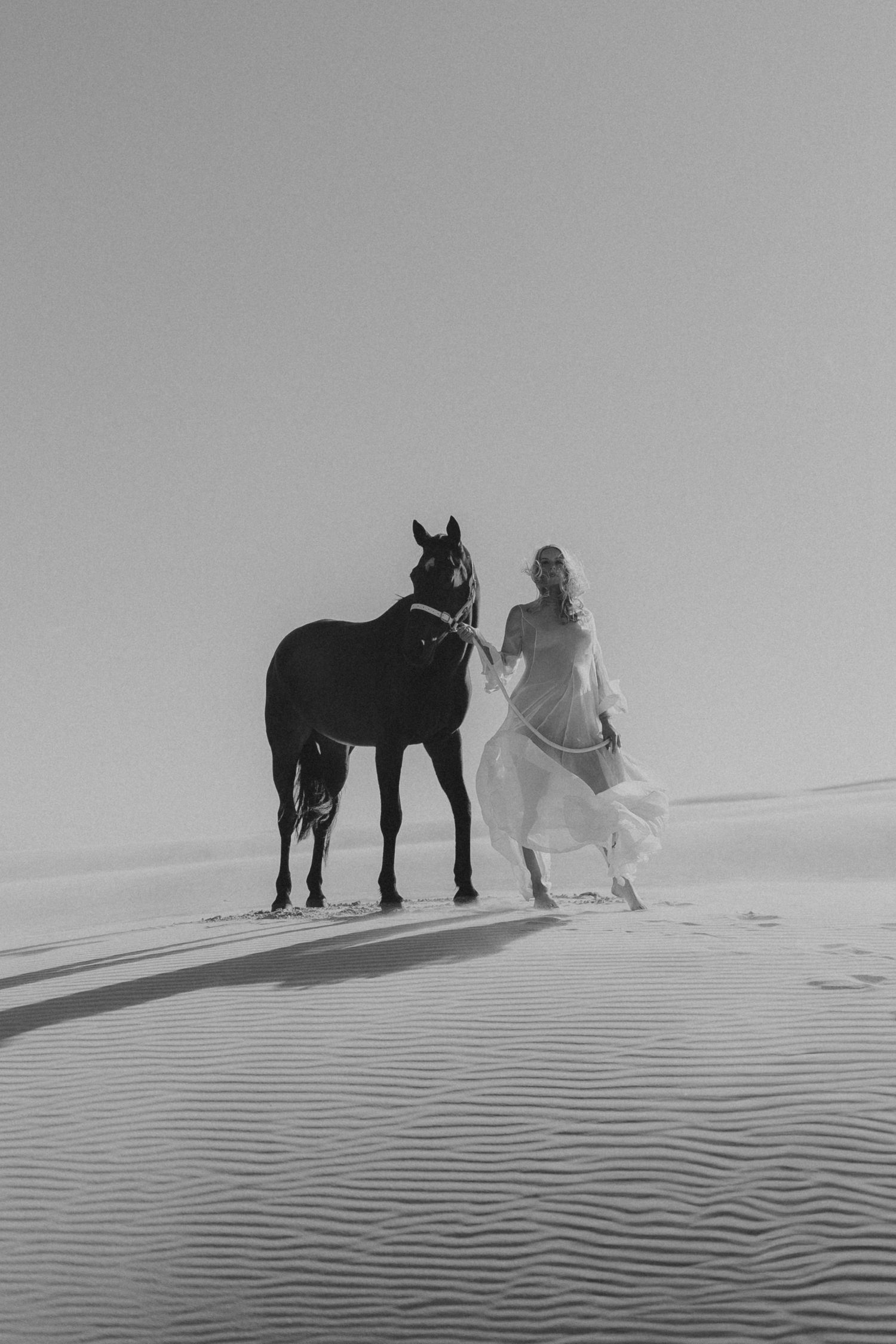 Black and white photo of a woman in a flowing white dress walking with a dark horse on sandy desert dunes.