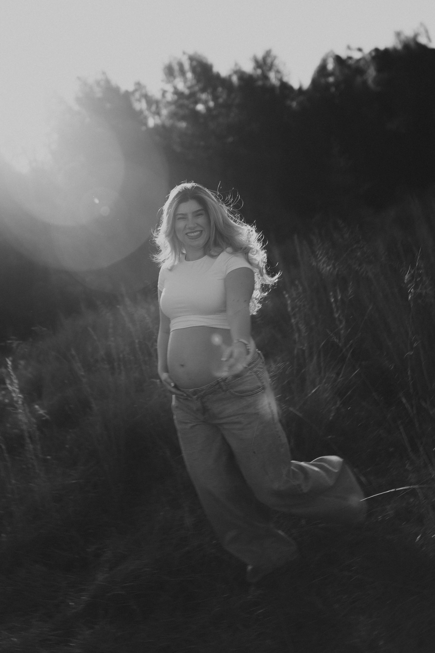 Black and white maternity photo of a smiling pregnant woman in a crop top and jeans in a sunlit field.