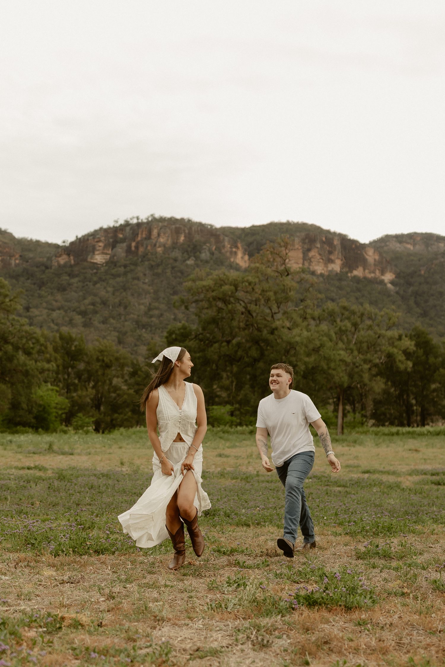 A couple runs playfully through a grassy field with rugged mountain cliffs rising in the distance.