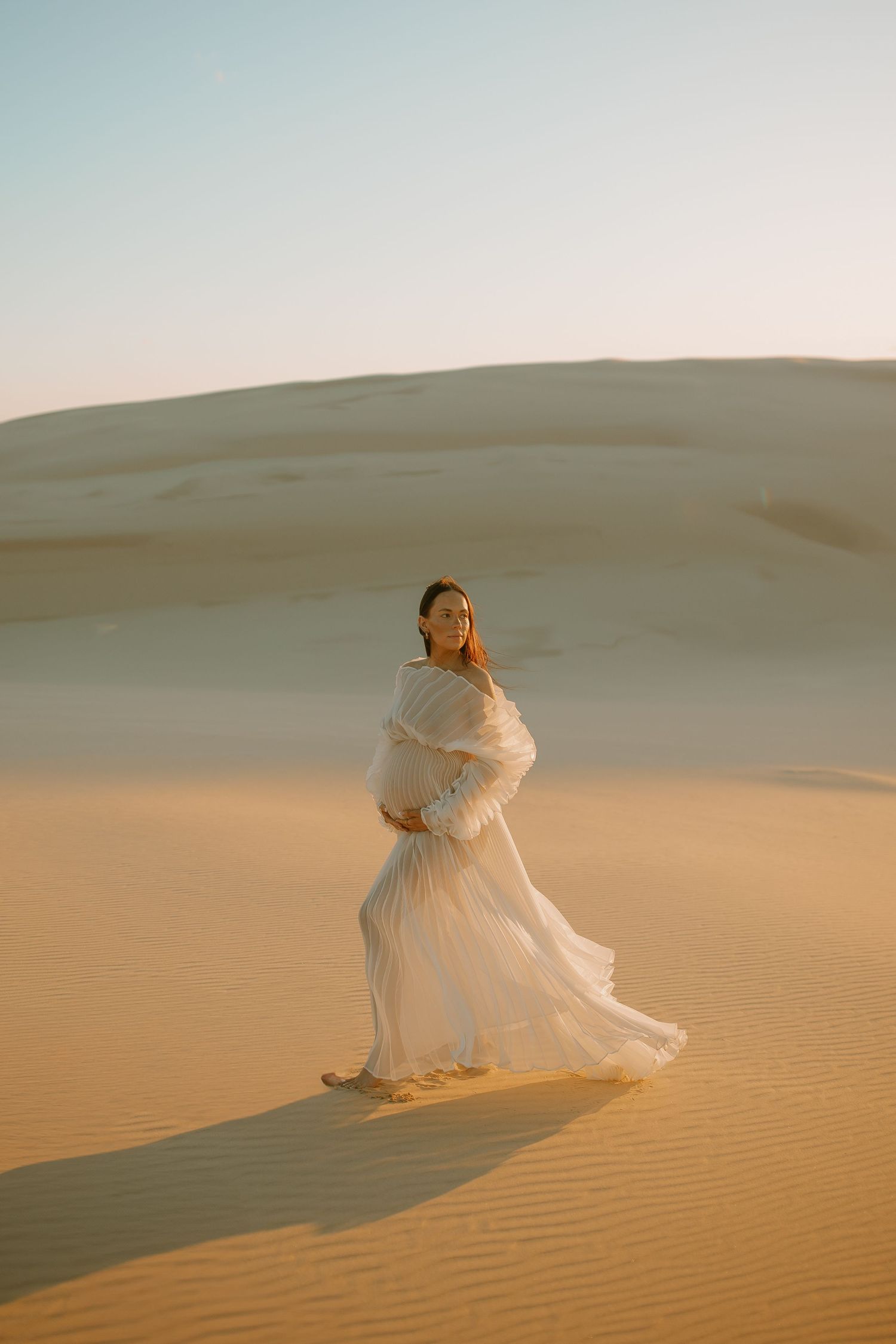 A figure in a flowing white dress sits gracefully atop golden sand dunes during sunset in a desert landscape.