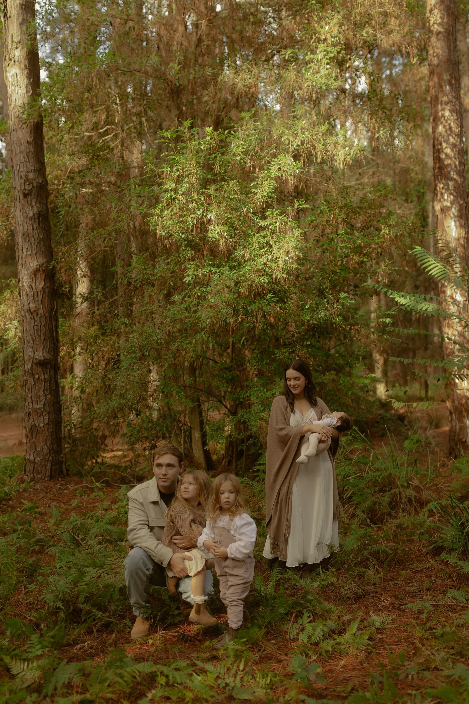 Family of four posing together in a forest setting with ferns and tall pine trees.