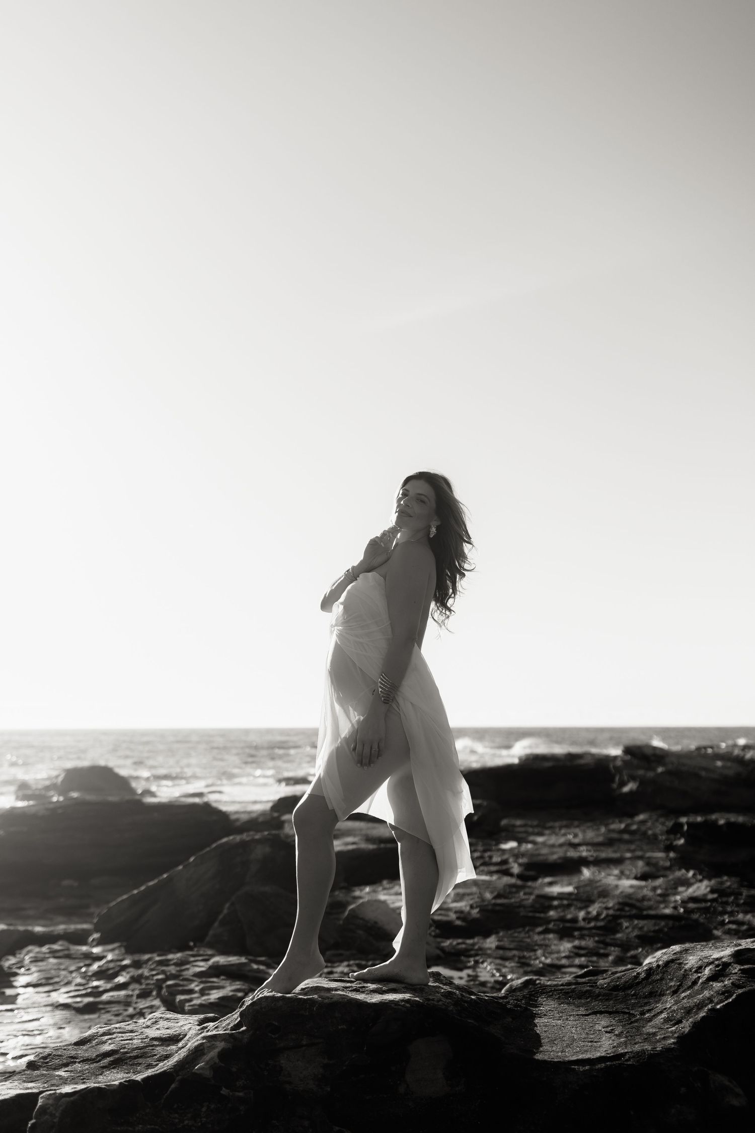 Black and white photo of a woman in a white dress standing on coastal rocks with ocean waves in the background.