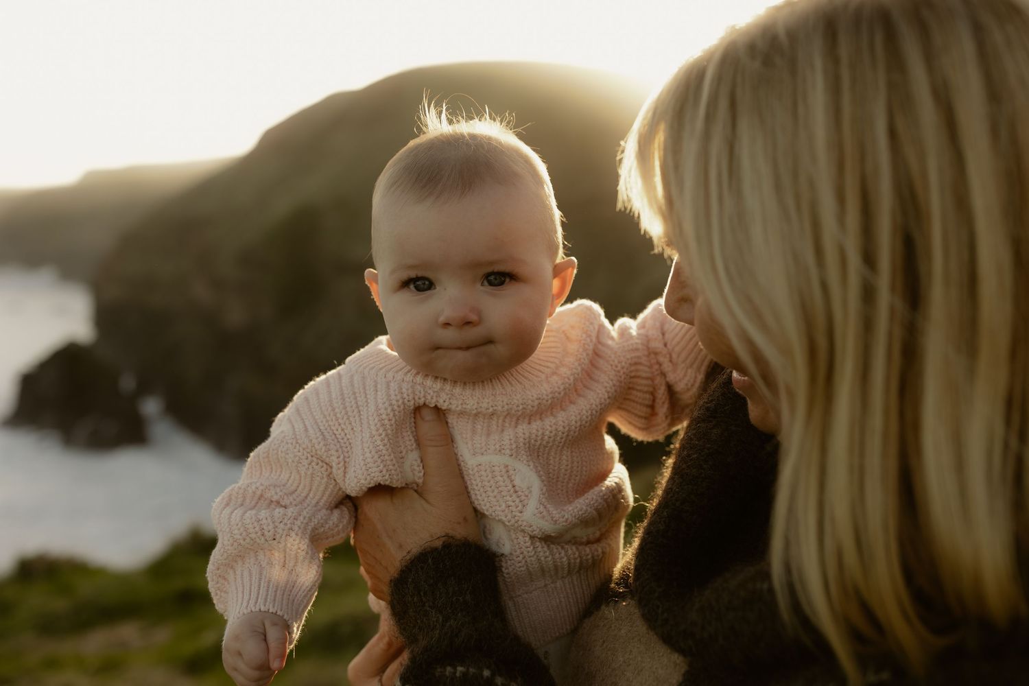 A mother holds her smiling baby in a pink knit sweater with a scenic coastal backdrop at sunset.