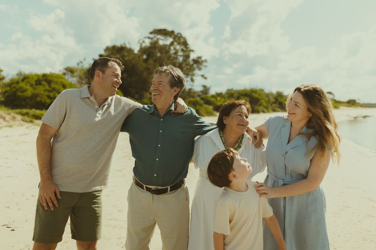 A group shares joyful laughter together on a sunny beach with trees in the background.