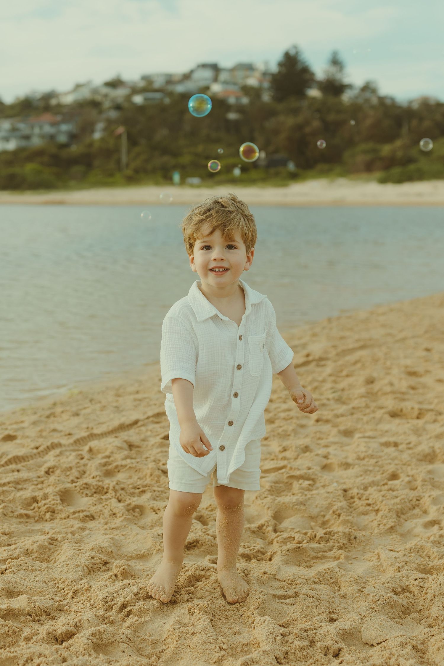 A child plays with bubbles on a sandy beach with water and houses in the background.