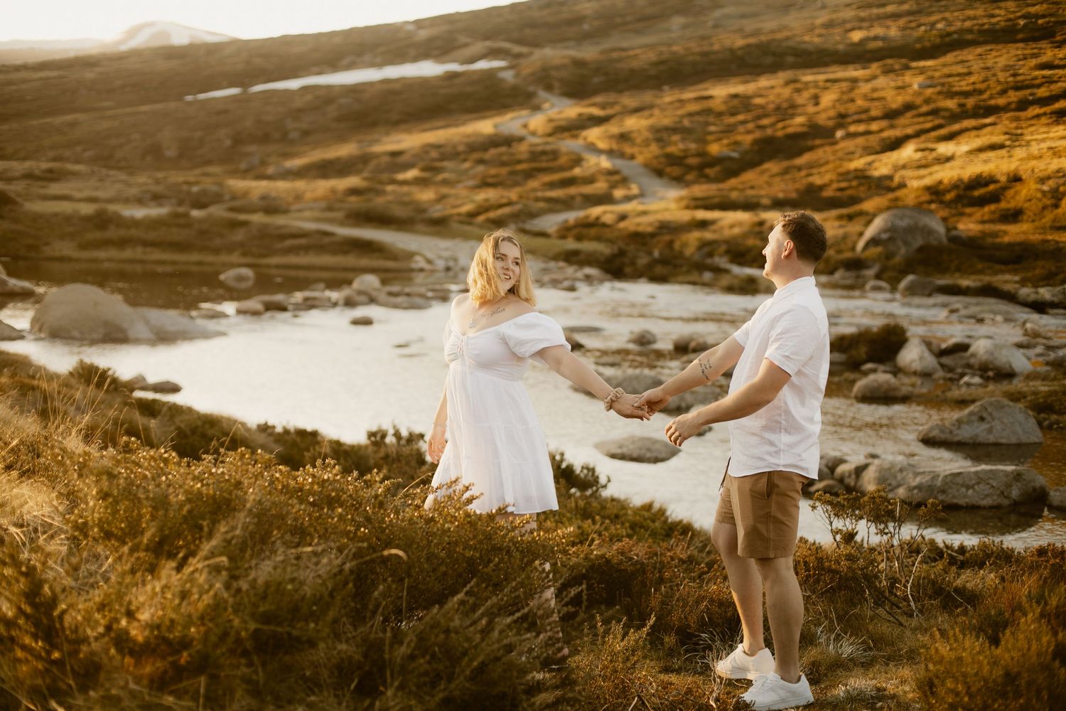 A couple in white outfits shares an intimate moment at sunset on a rugged coastal cliff overlooking the ocean.