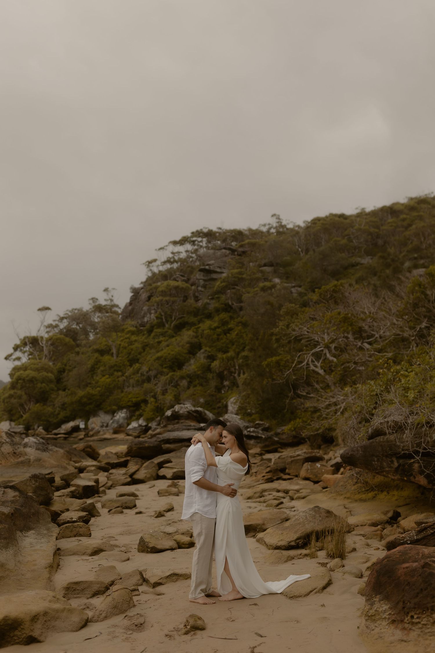 A romantic couple embraces on a rocky beach with lush green cliffs and overcast skies in the background.