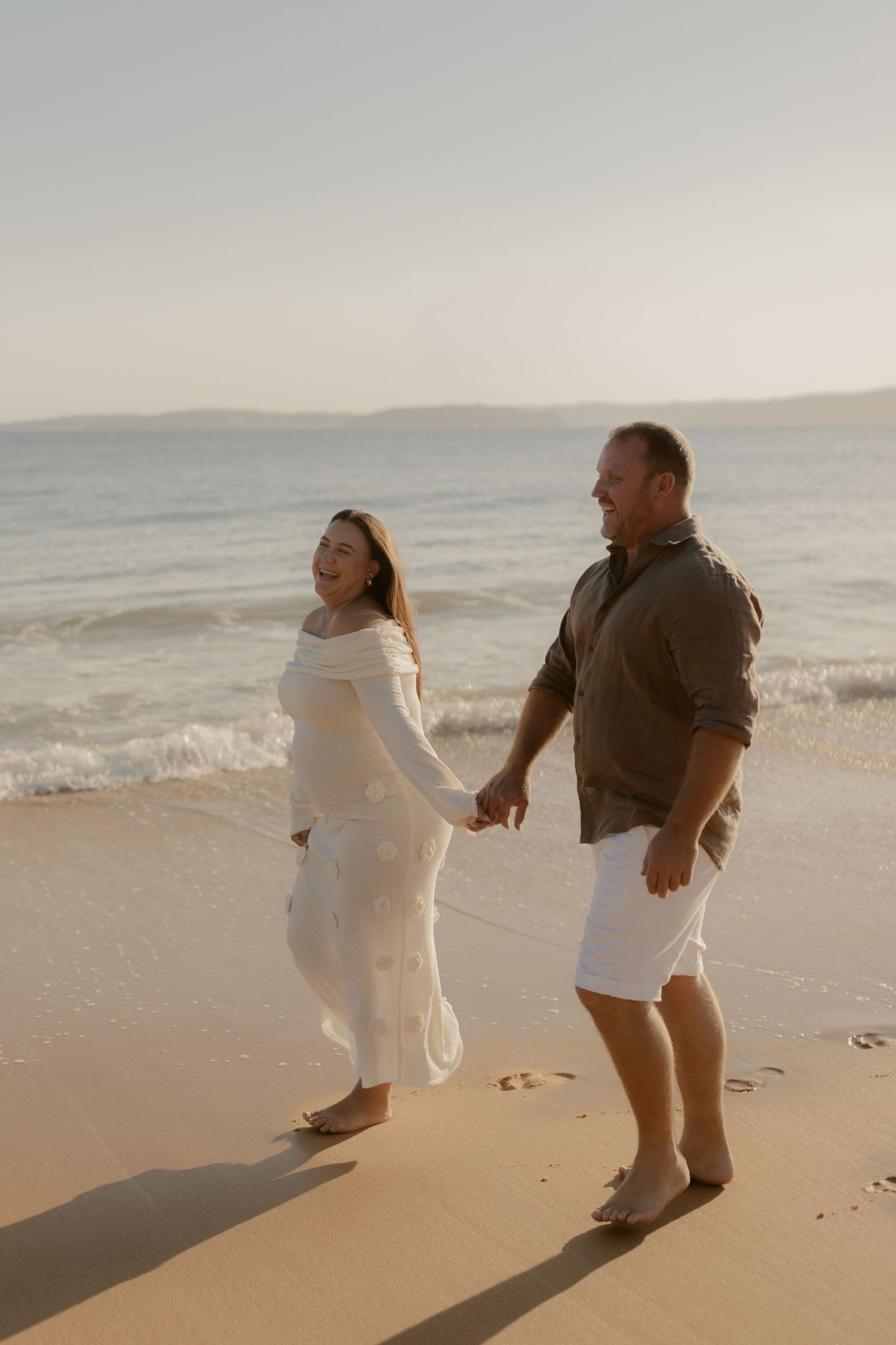 A couple walks hand in hand along a serene beach at sunset, creating long shadows in the sand.