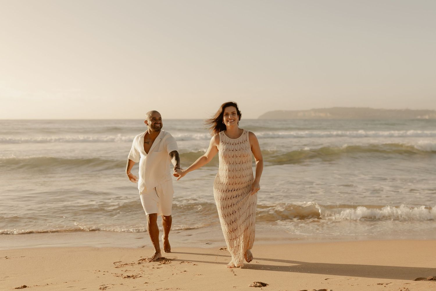 A couple in light colored dresses holds hands while walking together on a sandy beach at sunset.