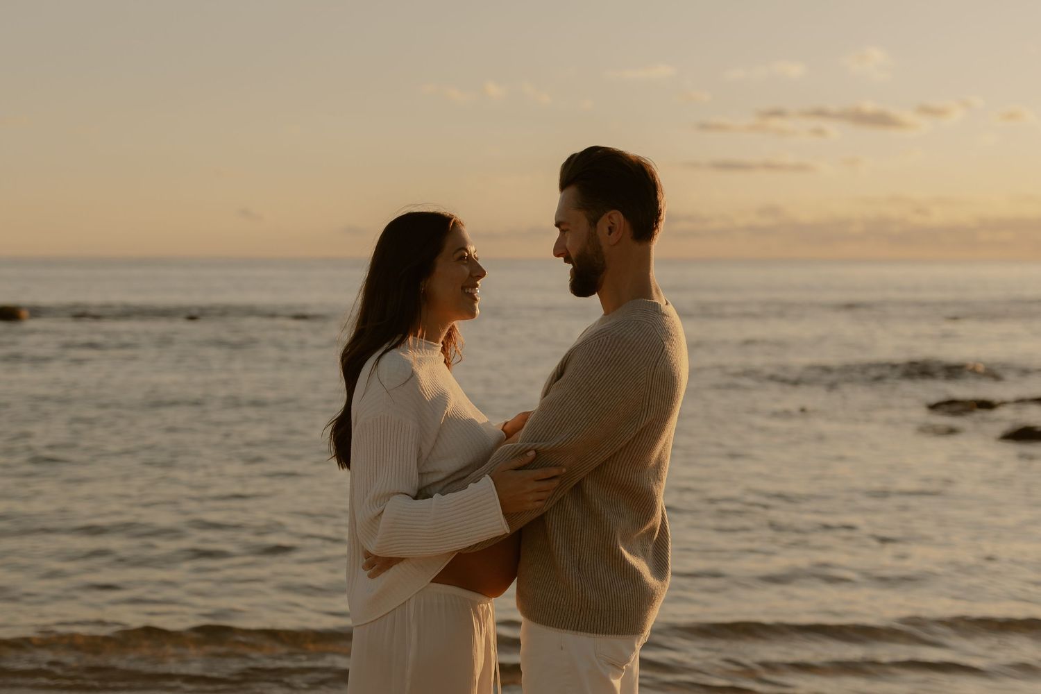 Romantic couple embraces on beach during golden sunset with waves crashing on shore in background.