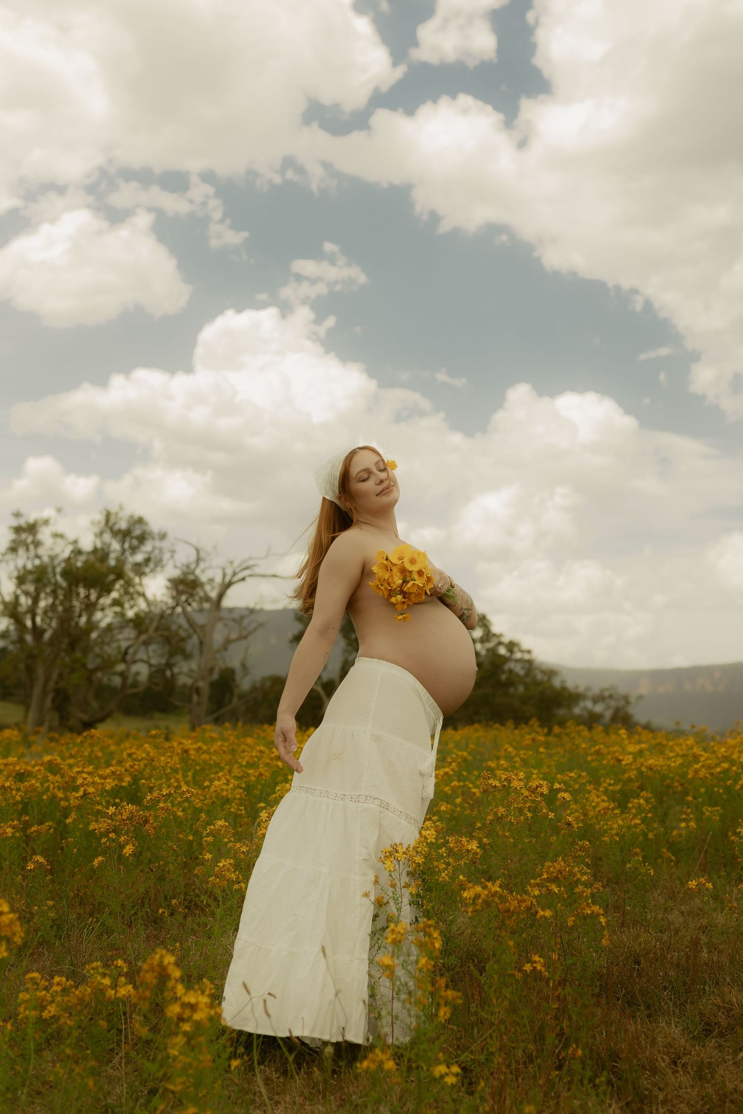 Pregnant woman in white skirt holding yellow flowers stands in a sunny field of wildflowers under a cloudy sky.