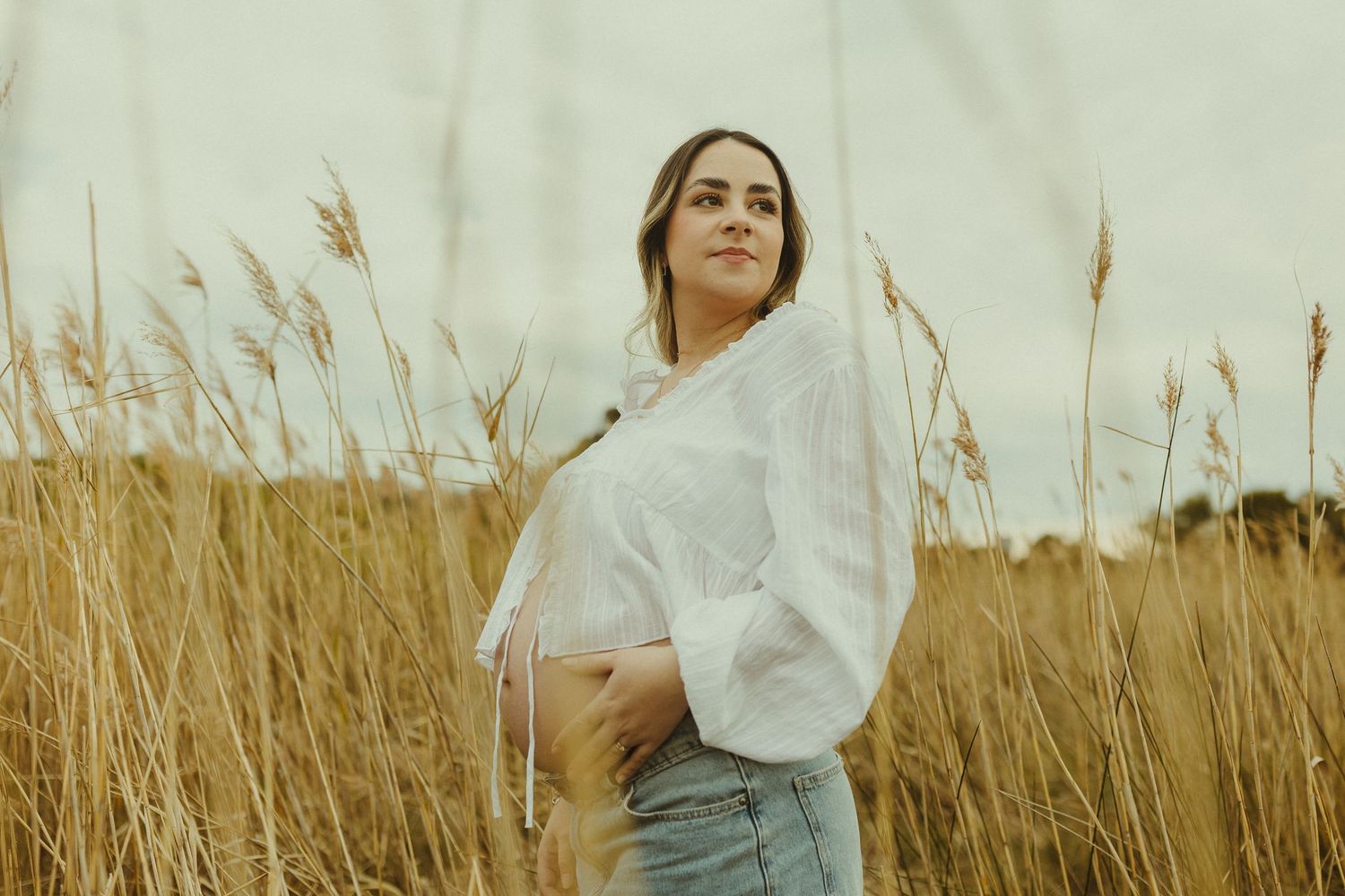Person poses joyfully in a golden wheat field at sunset wearing casual white clothing.