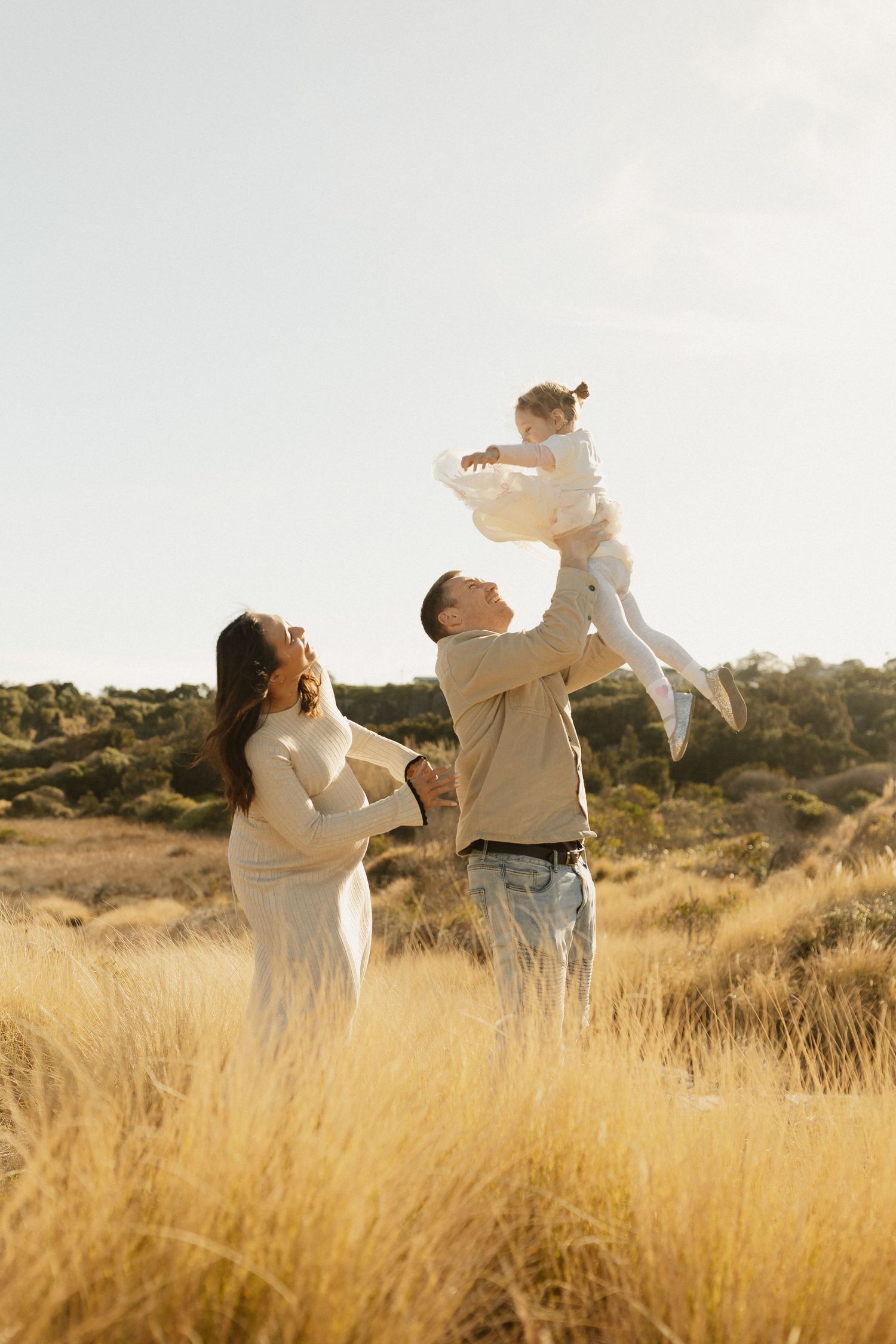 Family shares joyful moment lifting child in field of tall golden grass at sunset.