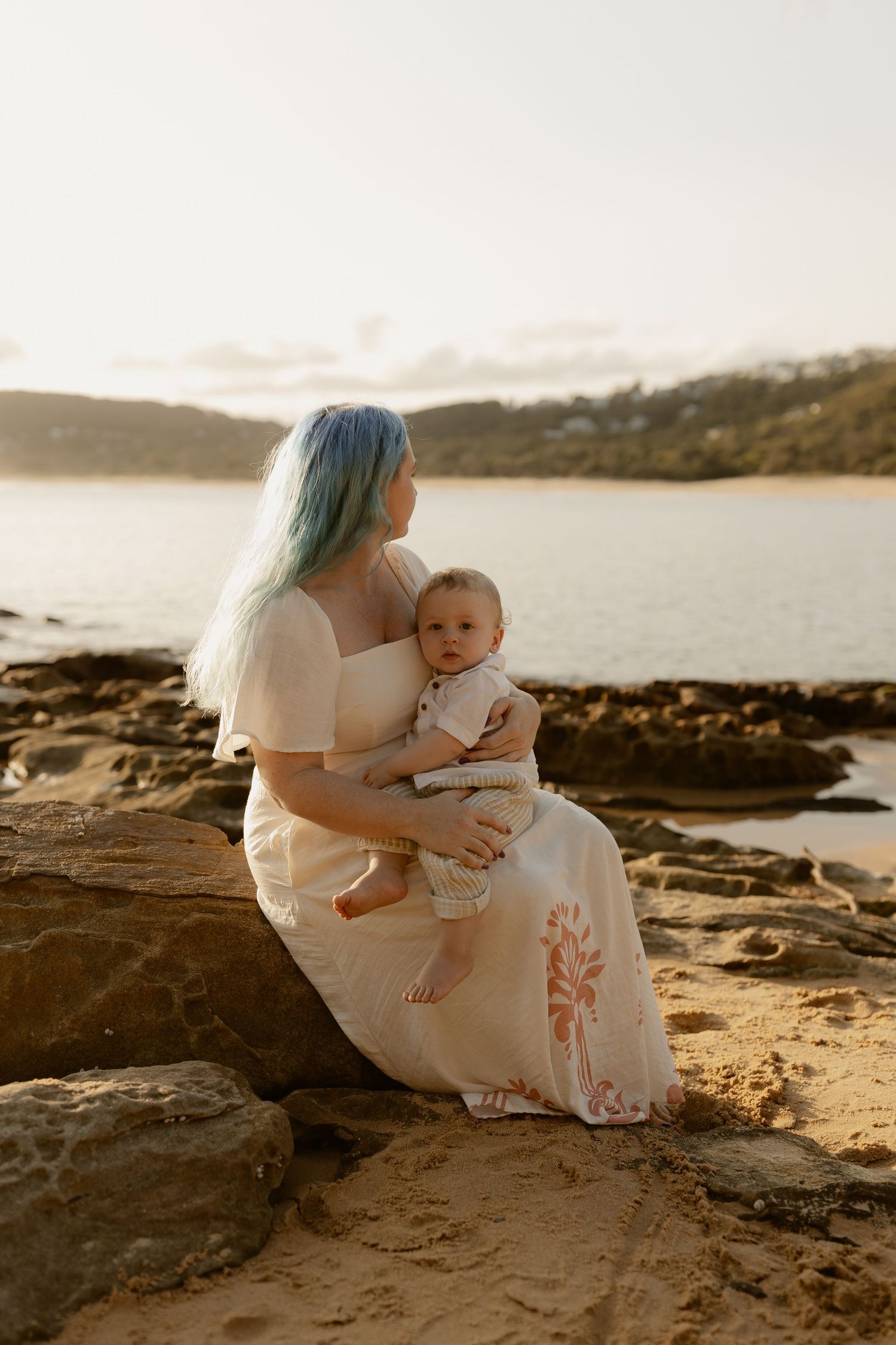Mother and child sitting together on coastal rocks during golden hour at a peaceful beach.
