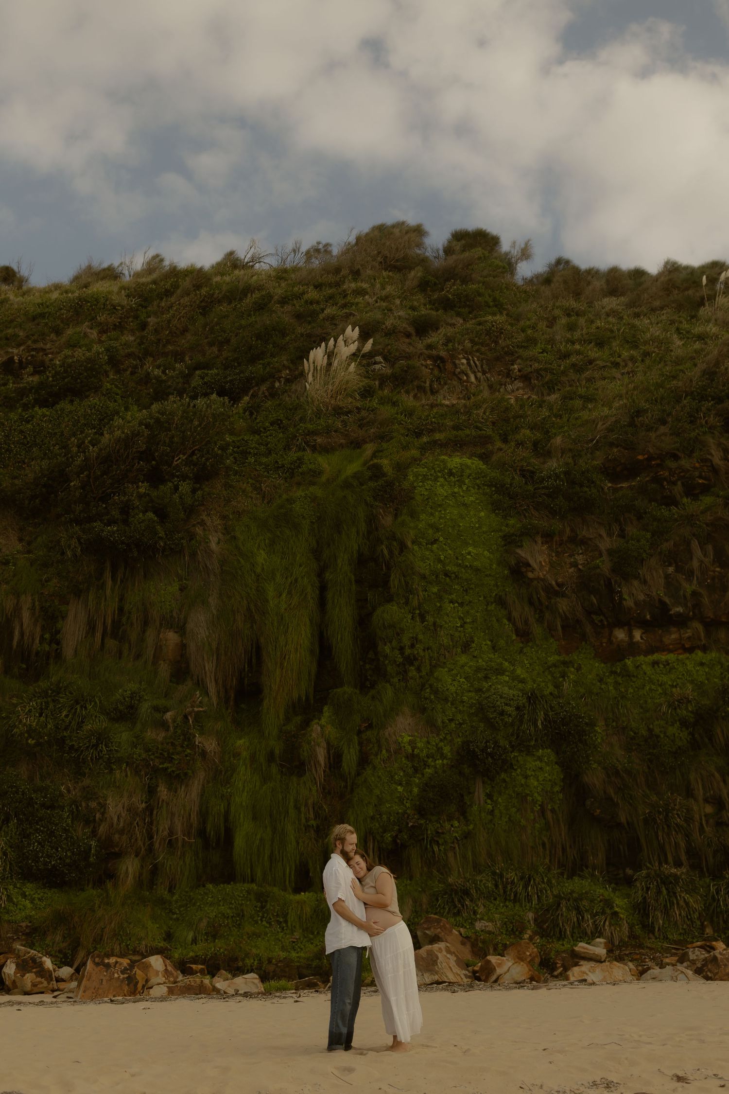 Couple stands together on a sandy beach with lush green cliffs in the background.