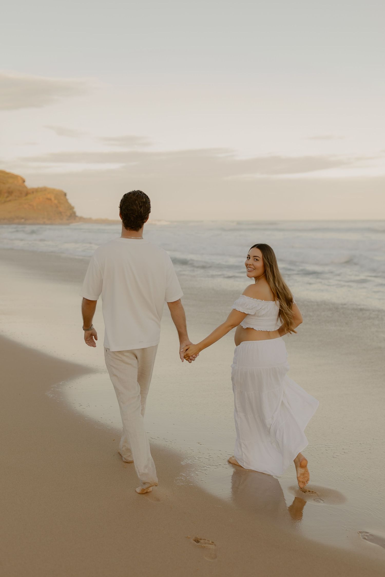 A couple in white outfits walking hand in hand along a scenic beach at sunset, creating romantic silhouettes against the ocean.