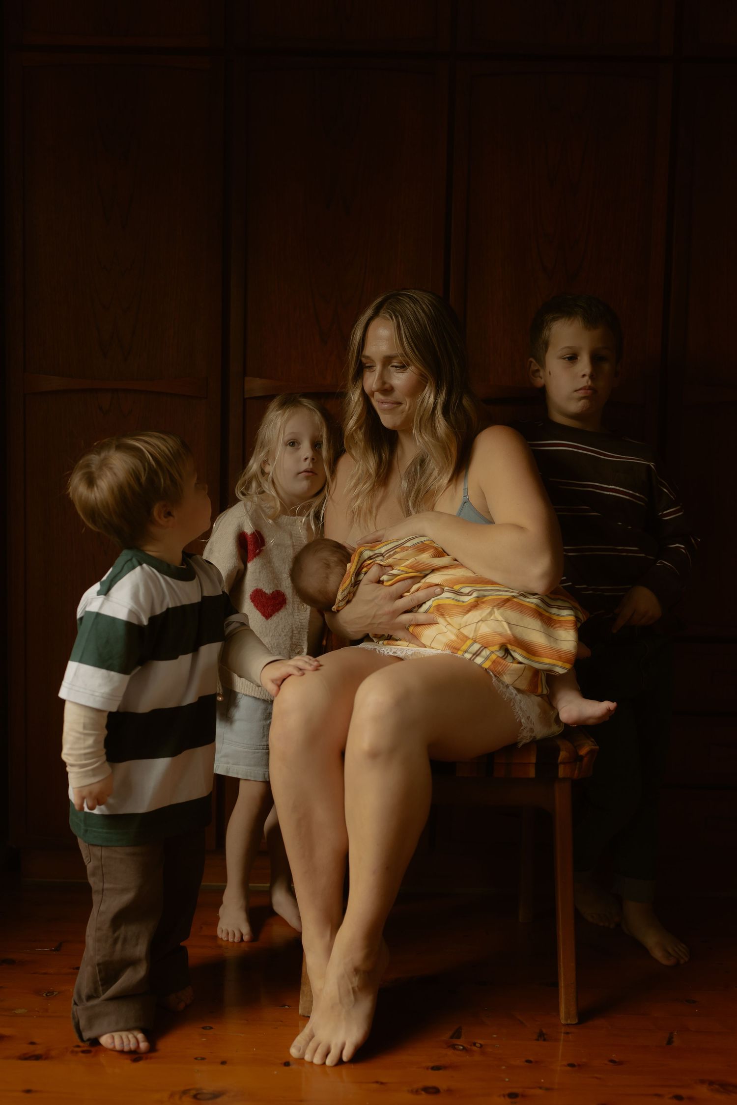 Mother sits barefoot holding newborn baby surrounded by three young children in a warmly lit room.