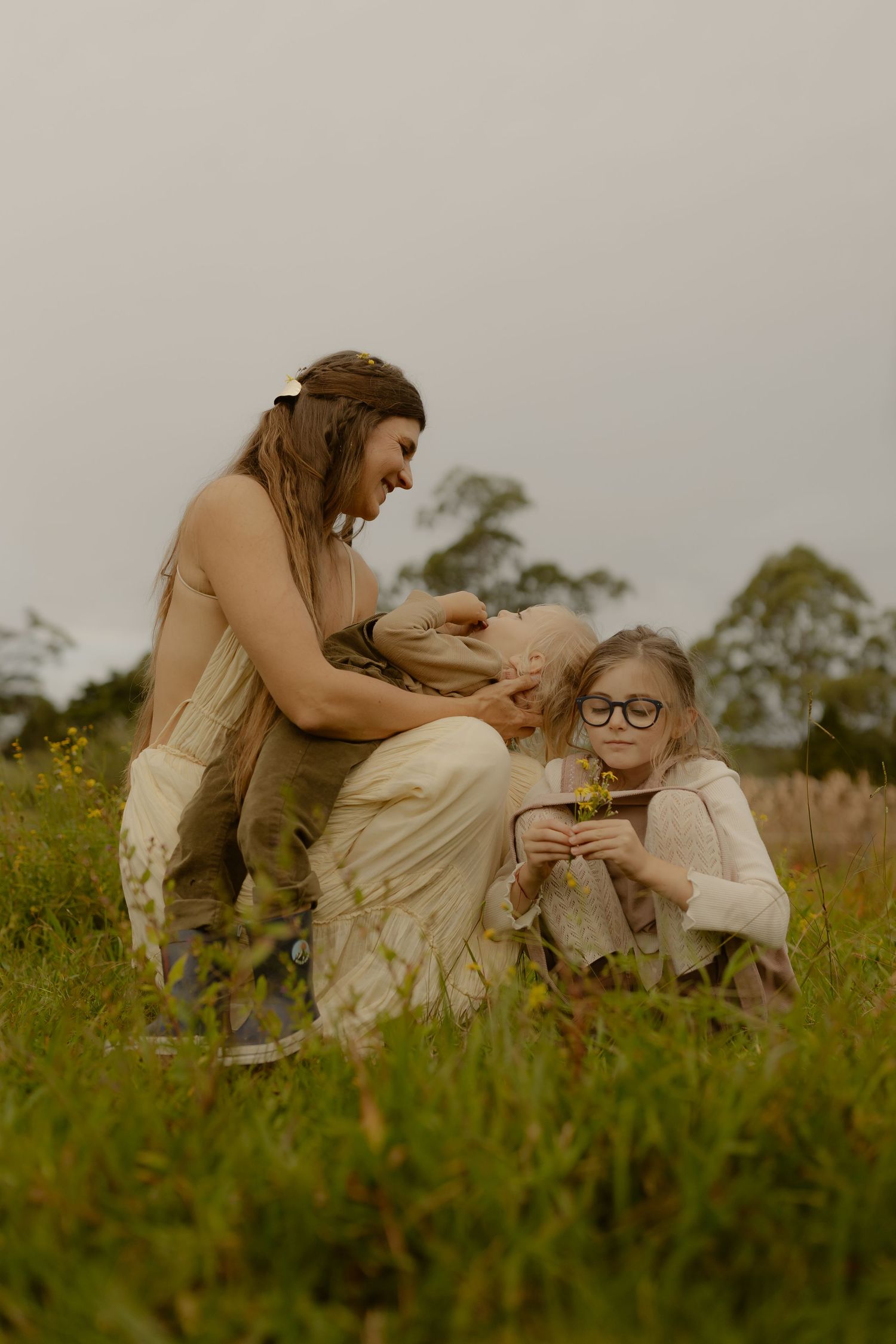 A mother and two children sit together in a green meadow picking wildflowers.