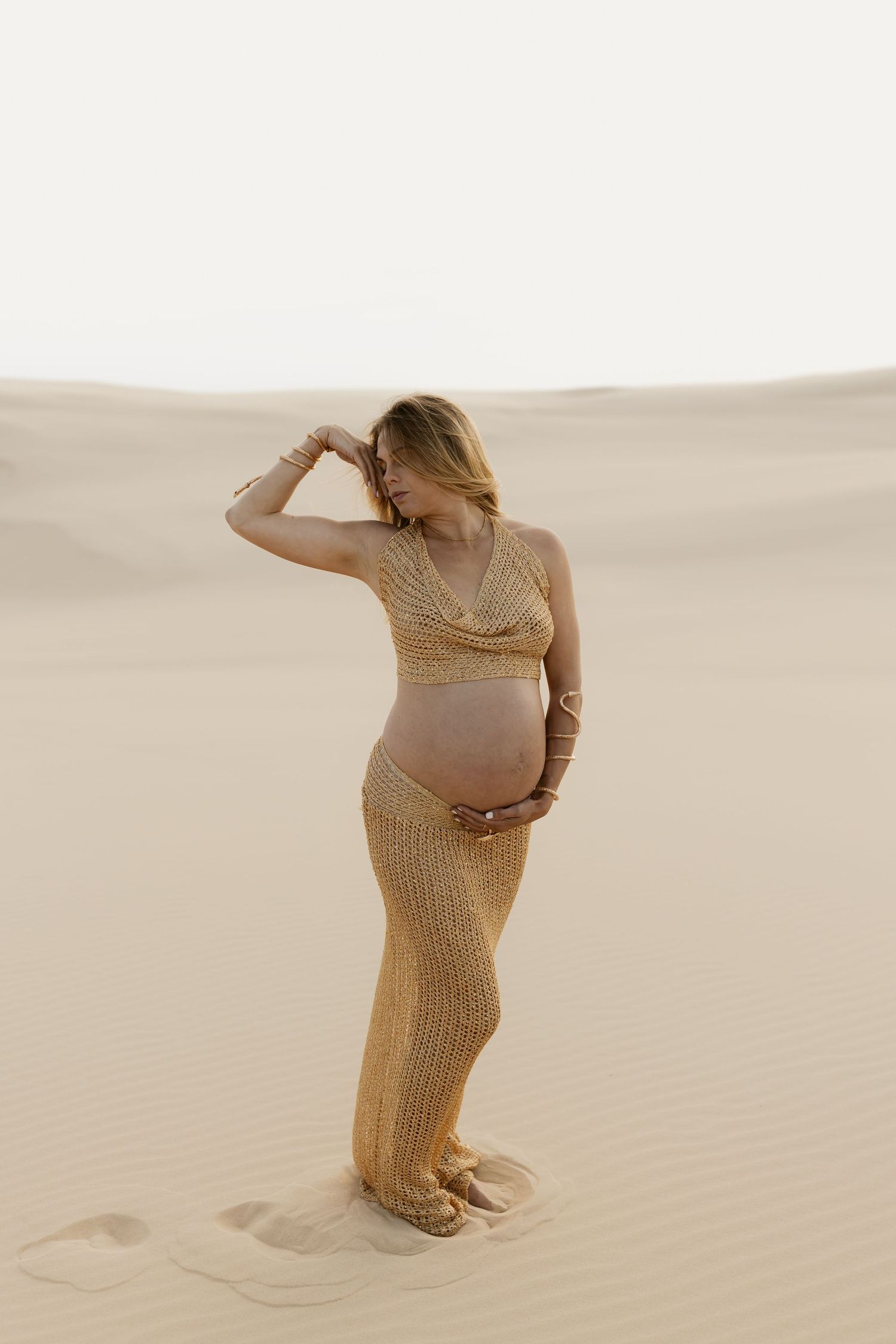 Maternity photoshoot in the desert with figure in golden dress posing against sandy dunes at sunset.