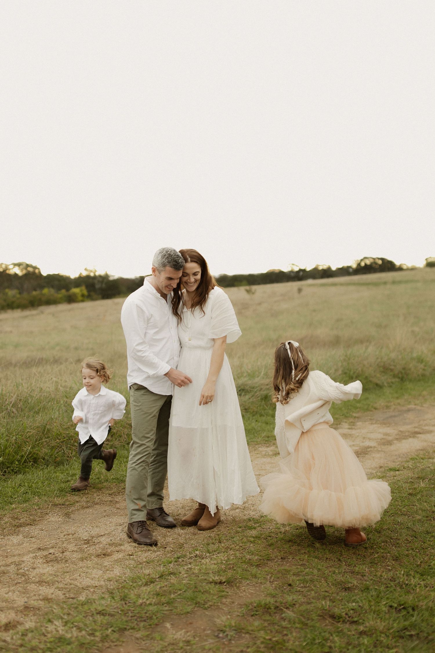 A playful scene unfolds on a dirt path through a grassy field as figures in white dresses dance and twirl together.
