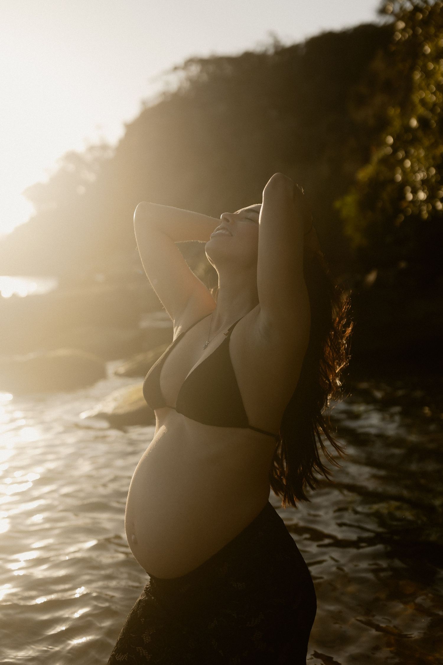 Maternity photoshoot at sunset with dramatic backlighting on a rocky beach.